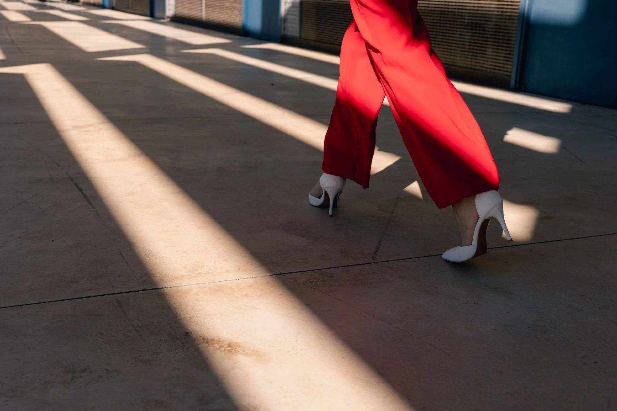 Piernas de una persona caminando con zapatos de tacón altos blancos, pantalones anchos de color rojo y fondo de sombra en la acera.