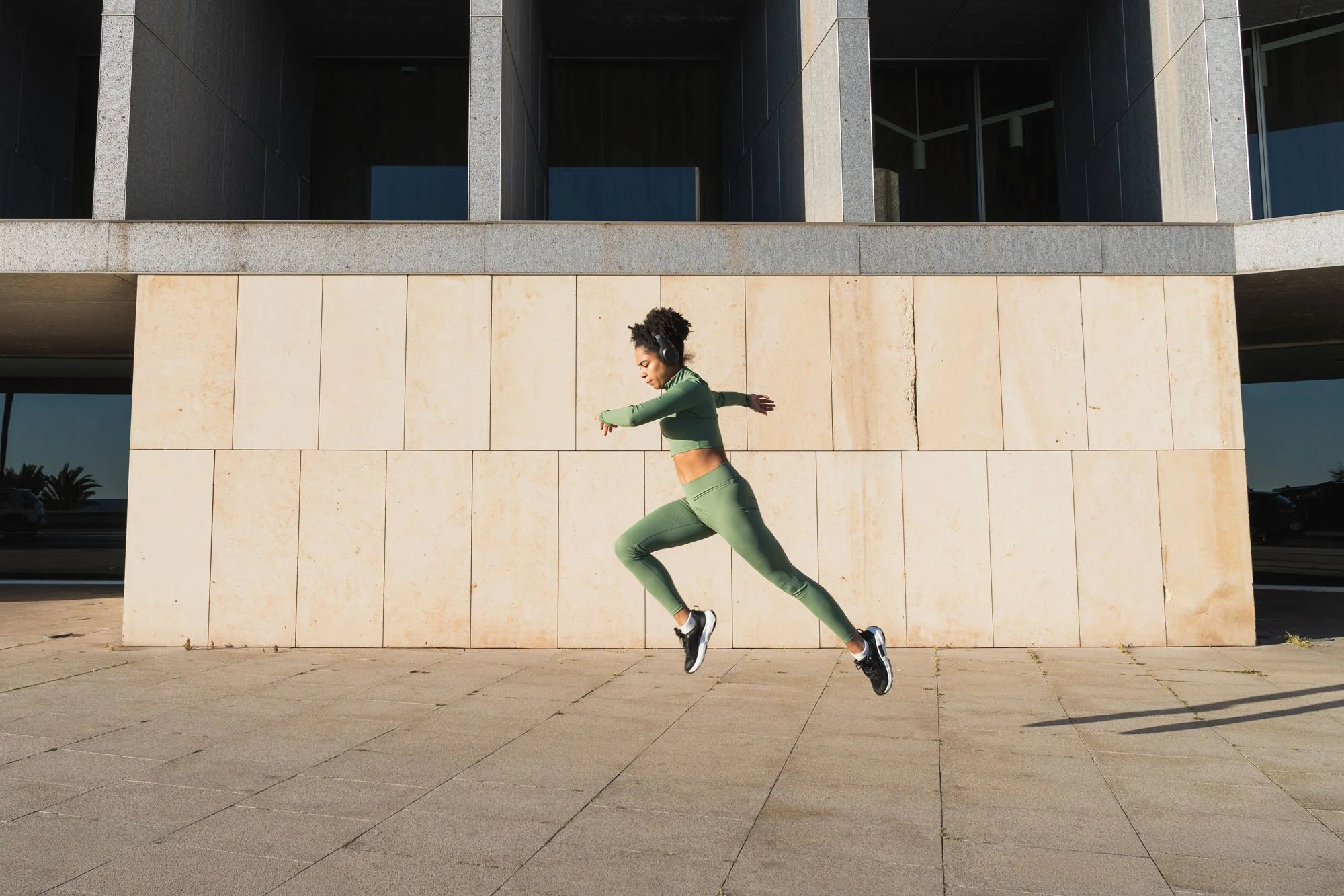 Mujer entrenando en movimientos de baile o ejercicio físico en un espacio urbano, usando ropa deportiva de color verde y auriculares, con un fondo de edificio moderno y cielo despejado.