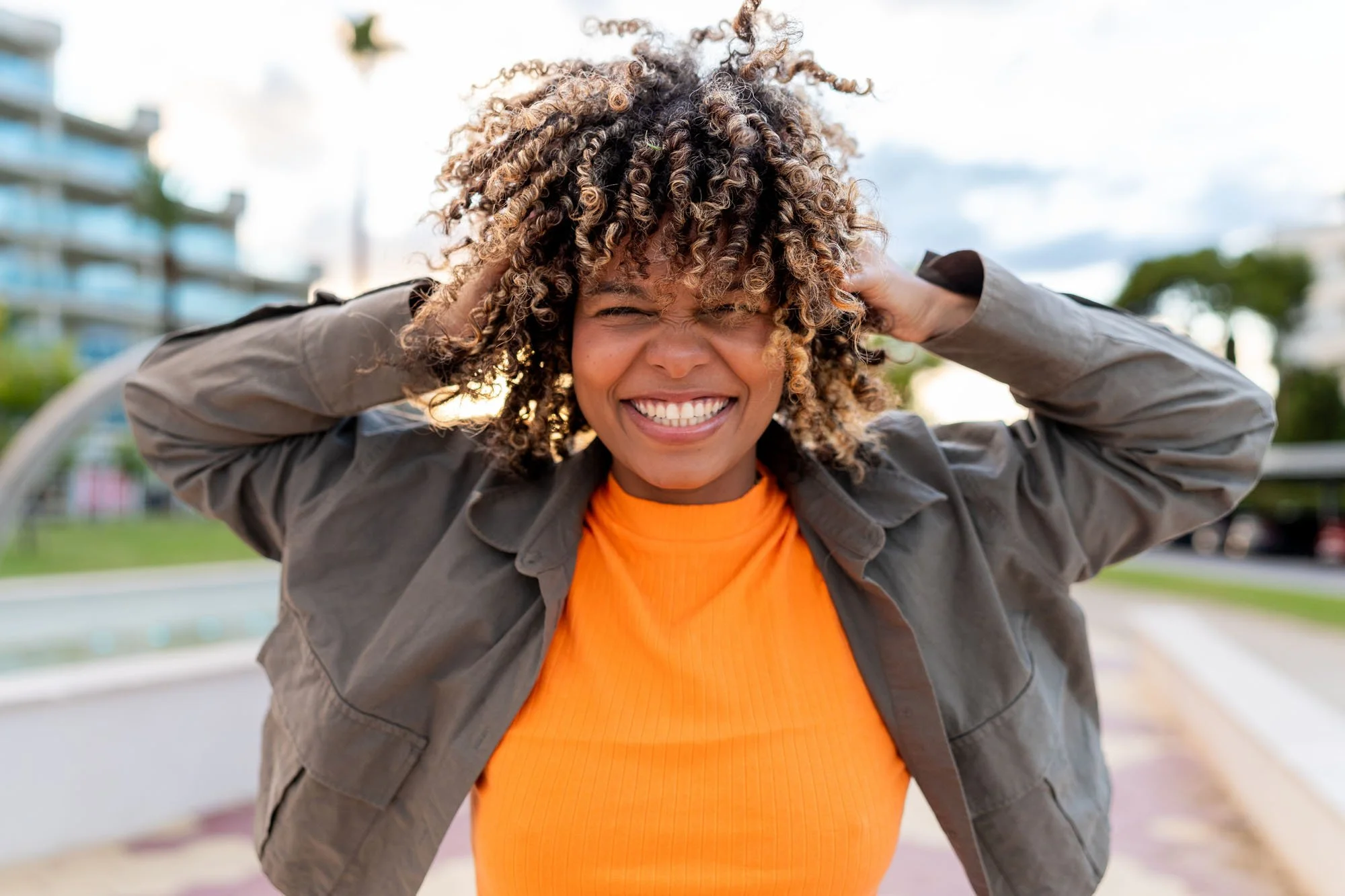 Mujer sonriente con cabello rizado, usando chaqueta gris y camiseta naranja, en un parque urbano.