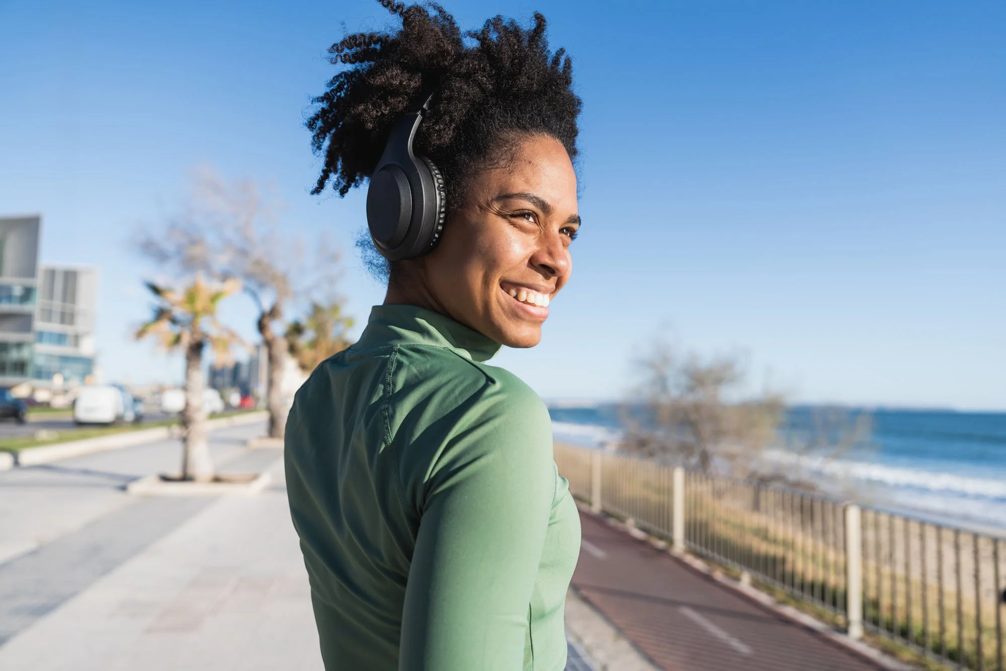 Mujer con peinado rizado usando audífonos grandes, sonriendo y mirando hacia la cámara, al aire libre junto a la playa con árboles y edificios a la distancia.