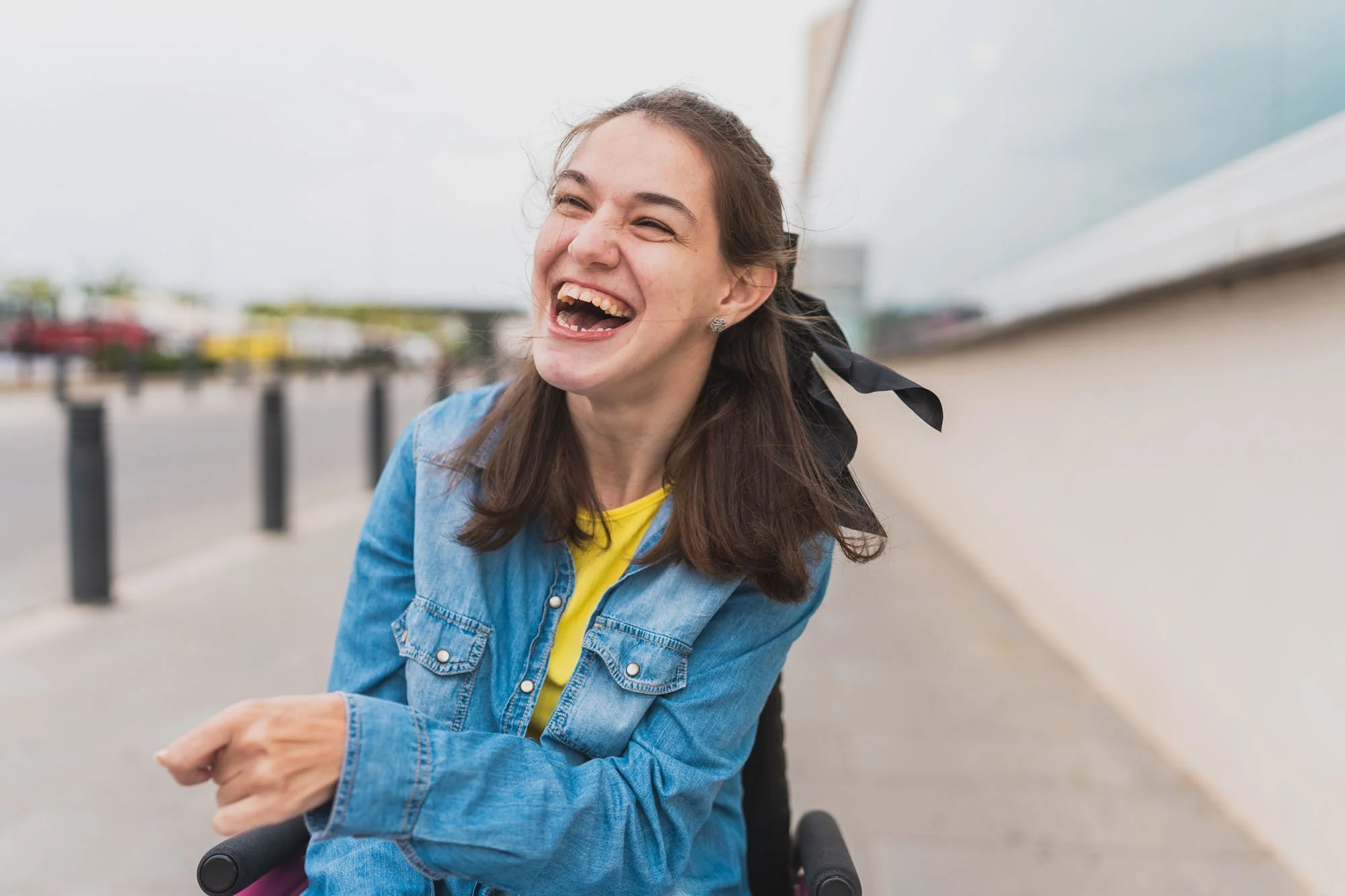 Joven mujer sonriendo y riendo en el exterior con fondo de parque o vía pública, usando chaqueta de mezclilla y camiseta amarilla.