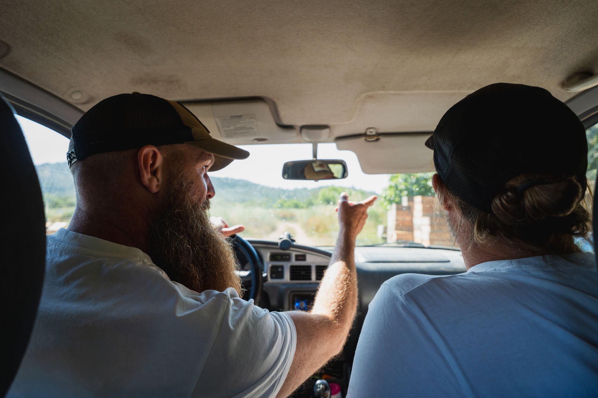Dos personas en el interior de un vehículo, una señalando algo con la mano, mientras conducen por un camino rural con vegetación y colinas al fondo.