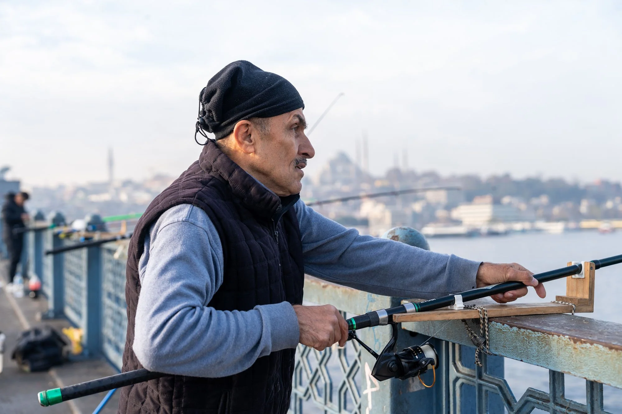 Hombre pescando con caña en un puente con vista de la ciudad al fondo, durante el día