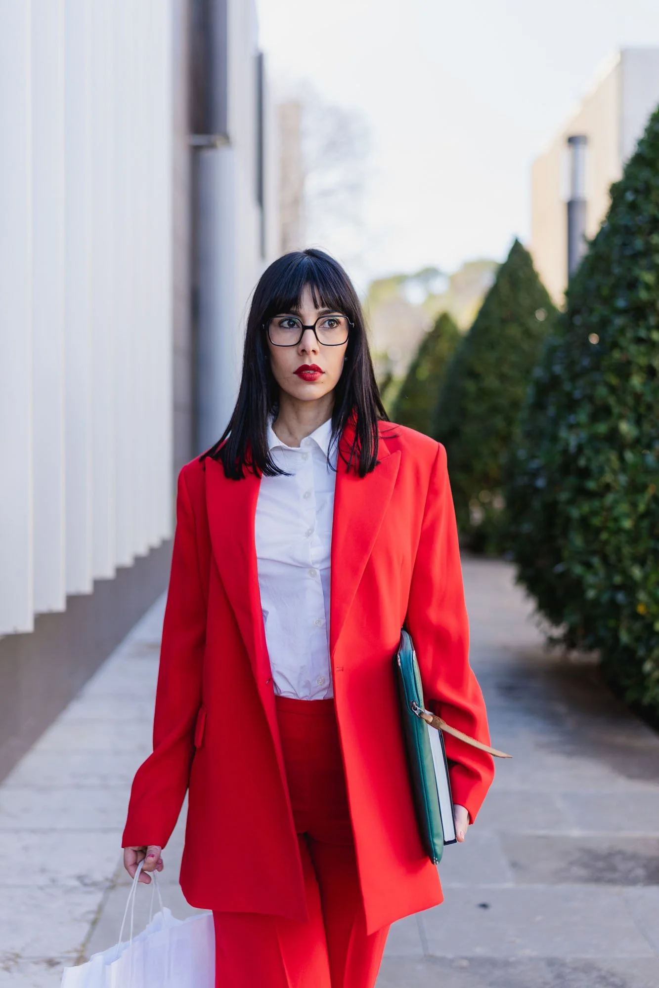 Mujer con gafas y cabello oscuro, vistiendo un traje rojo, caminando por una calle con arbustos verdes y edificios modernos al fondo, sosteniendo una bolsa blanca y un portafolio azul.