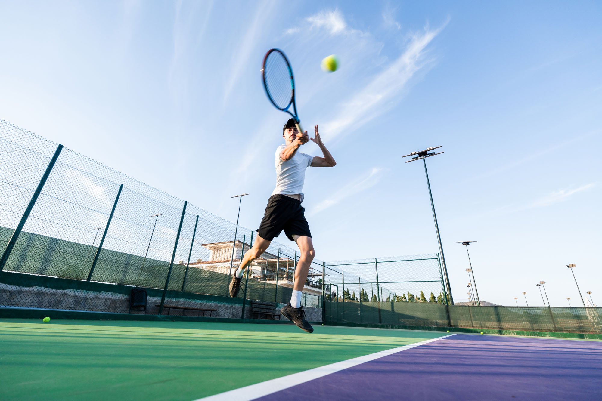 Hombre jugando tenis sobre cancha de color verde con red y vallas, lanzando una pelota con raqueta, bajo cielo despejado.