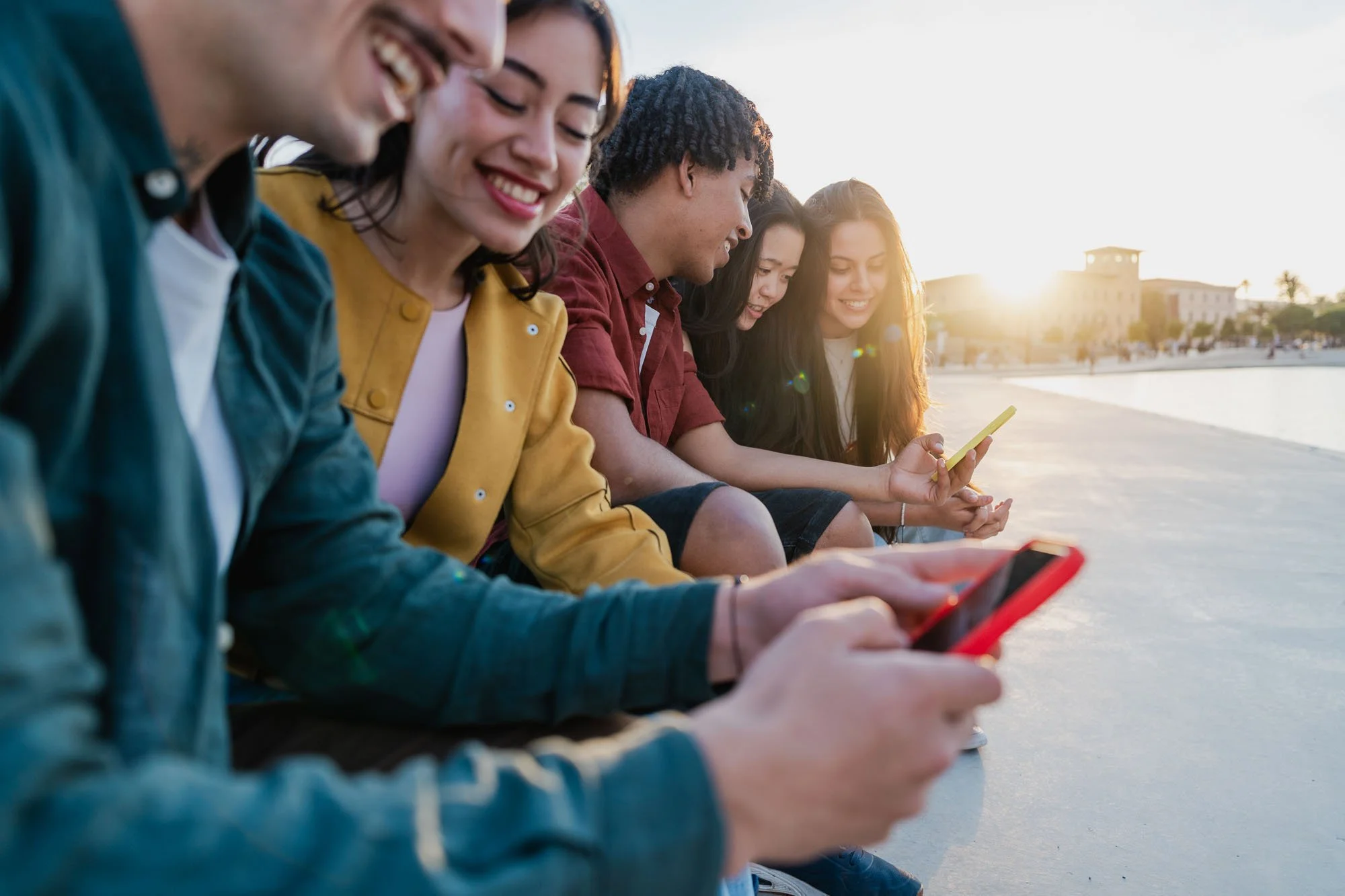 Grupo de jóvenes sentados en la orilla de un río o lago en la tarde, usando teléfonos móviles y sonriendo.
