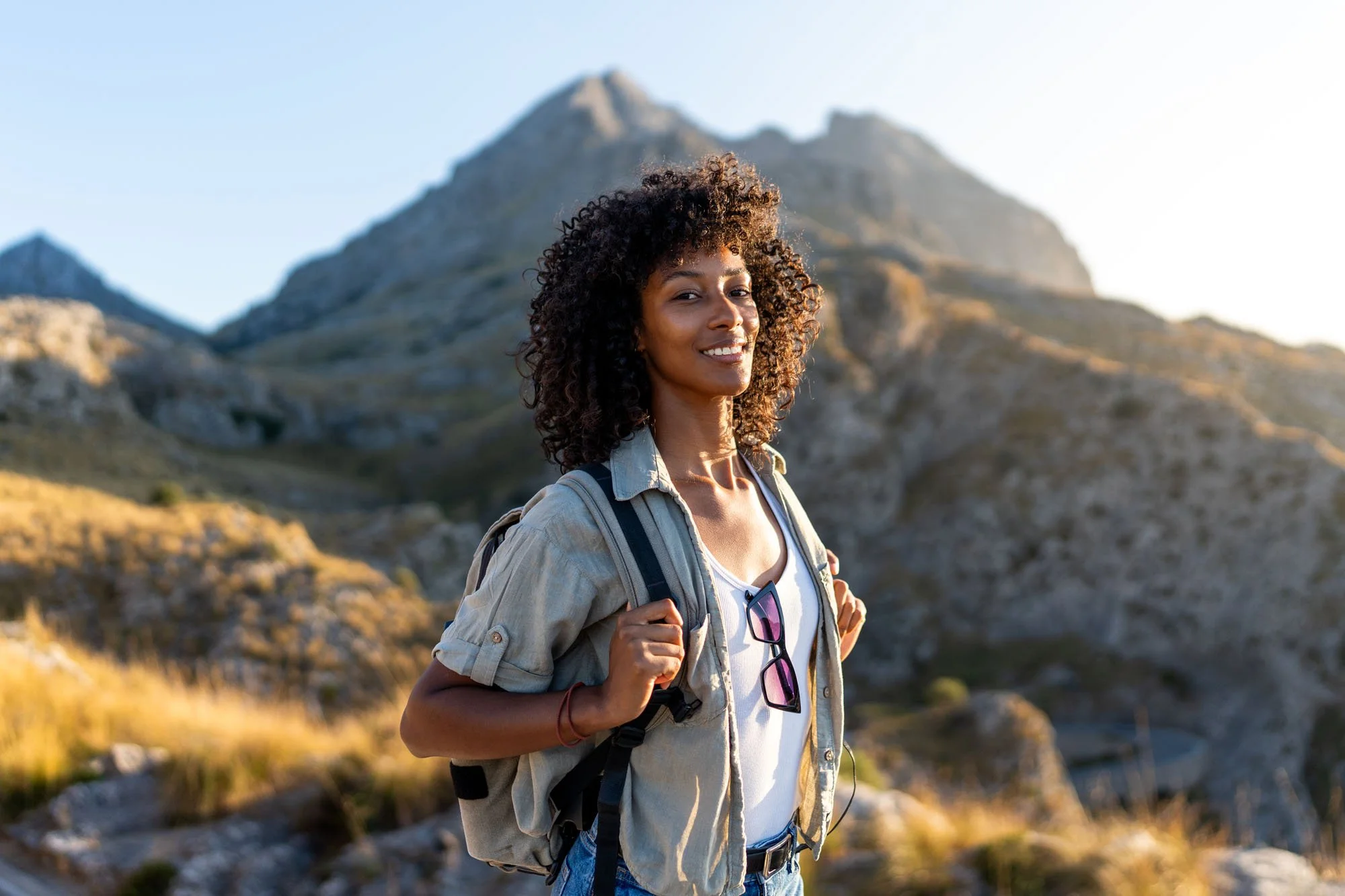 Mujer joven con mochila, camiseta blanca y gafas de sol colgando del cuello, caminando en un paisaje de montaña con cielo despejado.