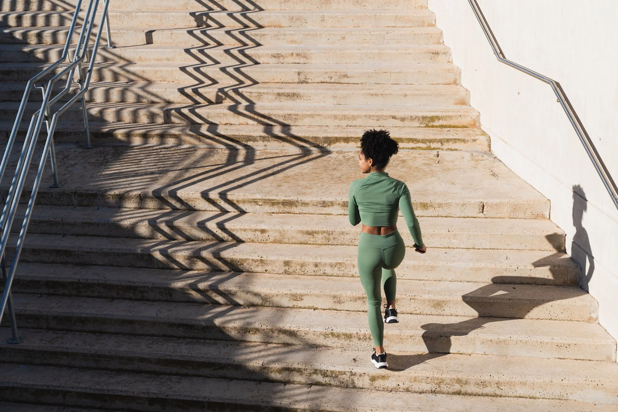 Mujer haciendo ejercicio subiendo unas escaleras de piedra, vestida con ropa deportiva verde, en un entorno al aire libre.