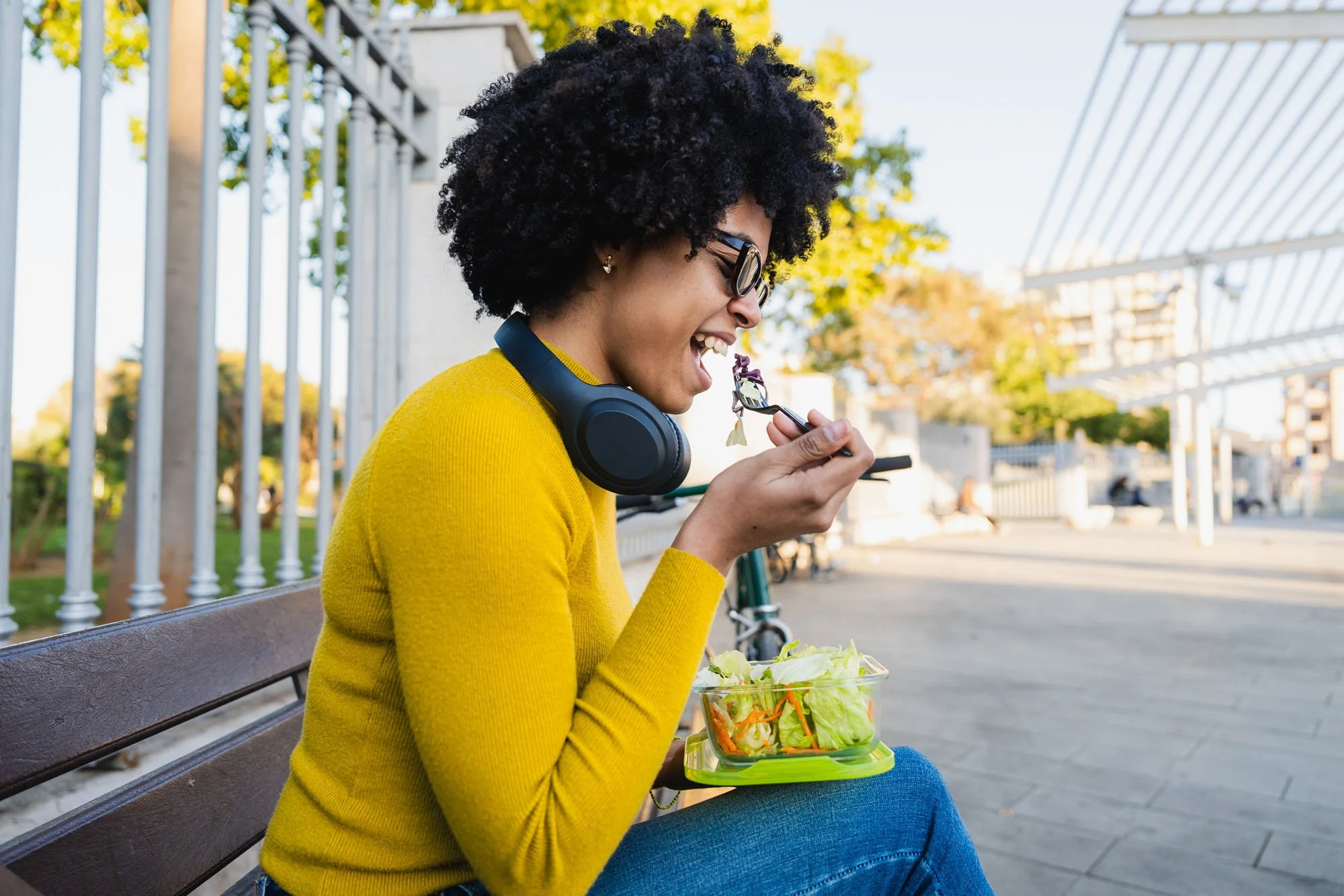Mujer sonriendo en un parque, comiendo ensalada y usando auriculares, con gafas y vestido amarillo.