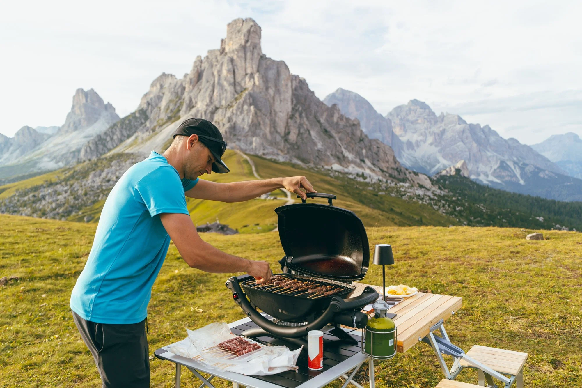 Un hombre que está cocinando carne en una parrilla portátil en un paisaje montañoso con césped y formaciones rocosas en el fondo.