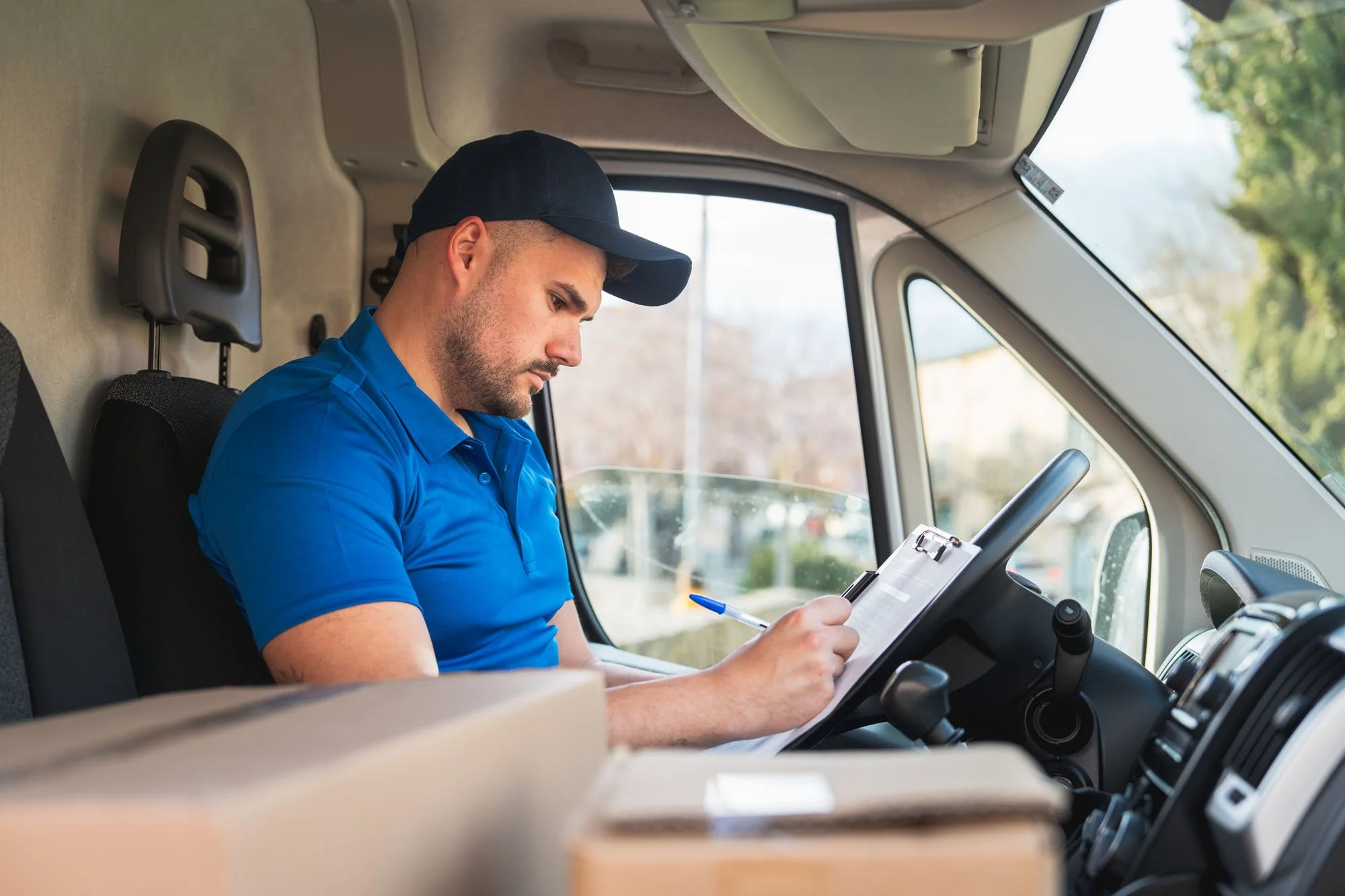 Un repartidor sentado en la cabina de un vehículo, usando una gorra y camisa azul, revisando un correo o recibiendo instrucciones en una libreta, con el interior del vehículo y detalles del exterior visible
