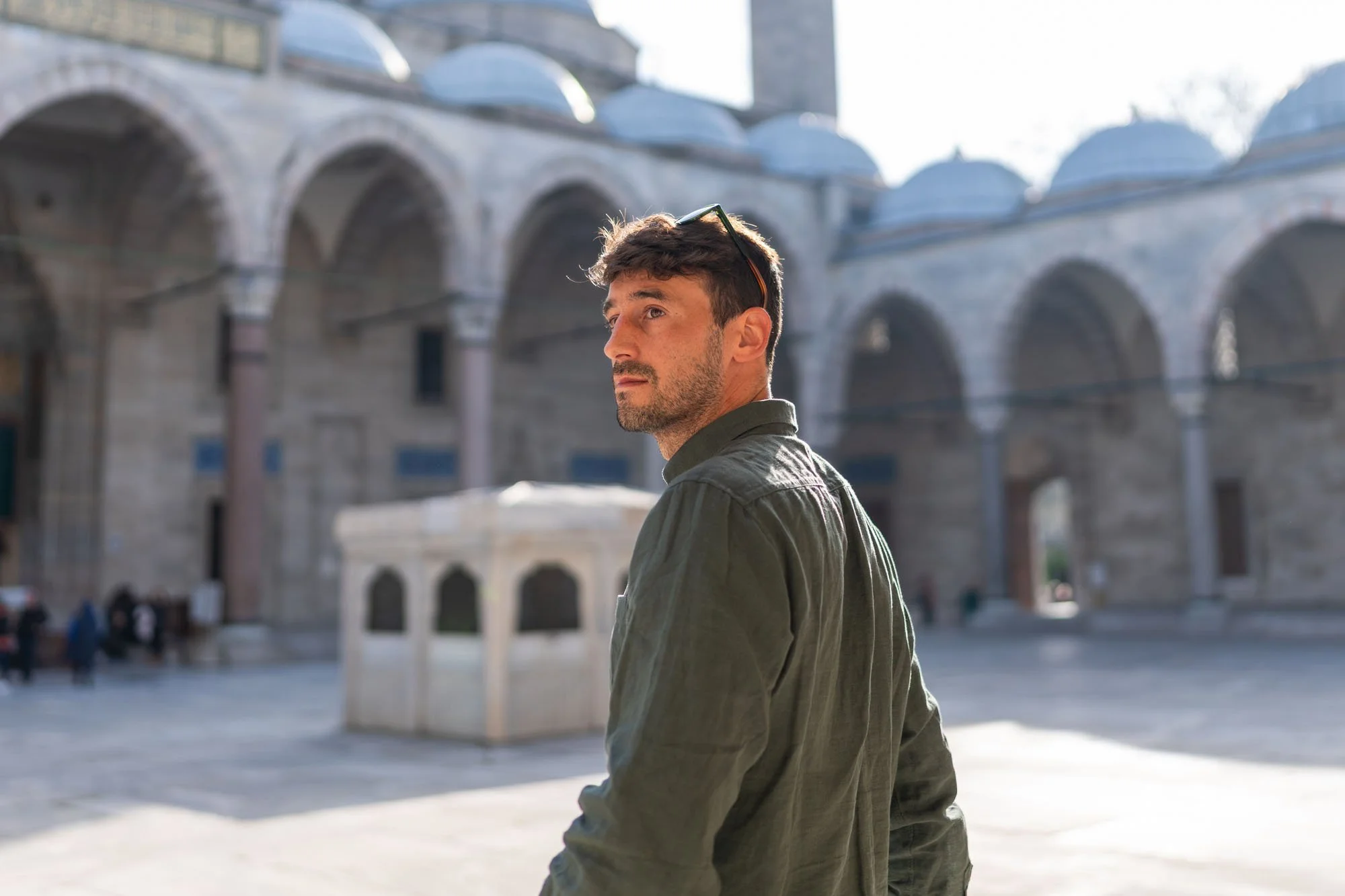 Joven hombre con barba y gafas sobre la cabeza, de perfil y expresión pensativa, de pie frente a la Basílica de Santa Sofía en Estambul, Turquía, con arquitectura otomana en el fondo y algunos turistas caminando alrededor.