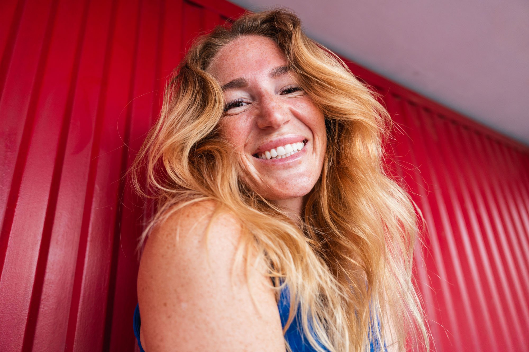 Mujer de cabello enrulado y rubio sonriendo sonriente, con un fondo de paredes rojas.