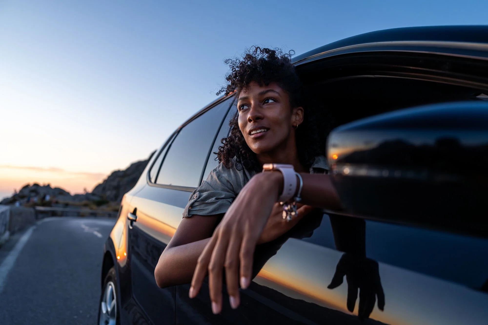 Una mujer con cabello rizado mira por la ventana de un automóvil negro durante el atardecer en una carretera.