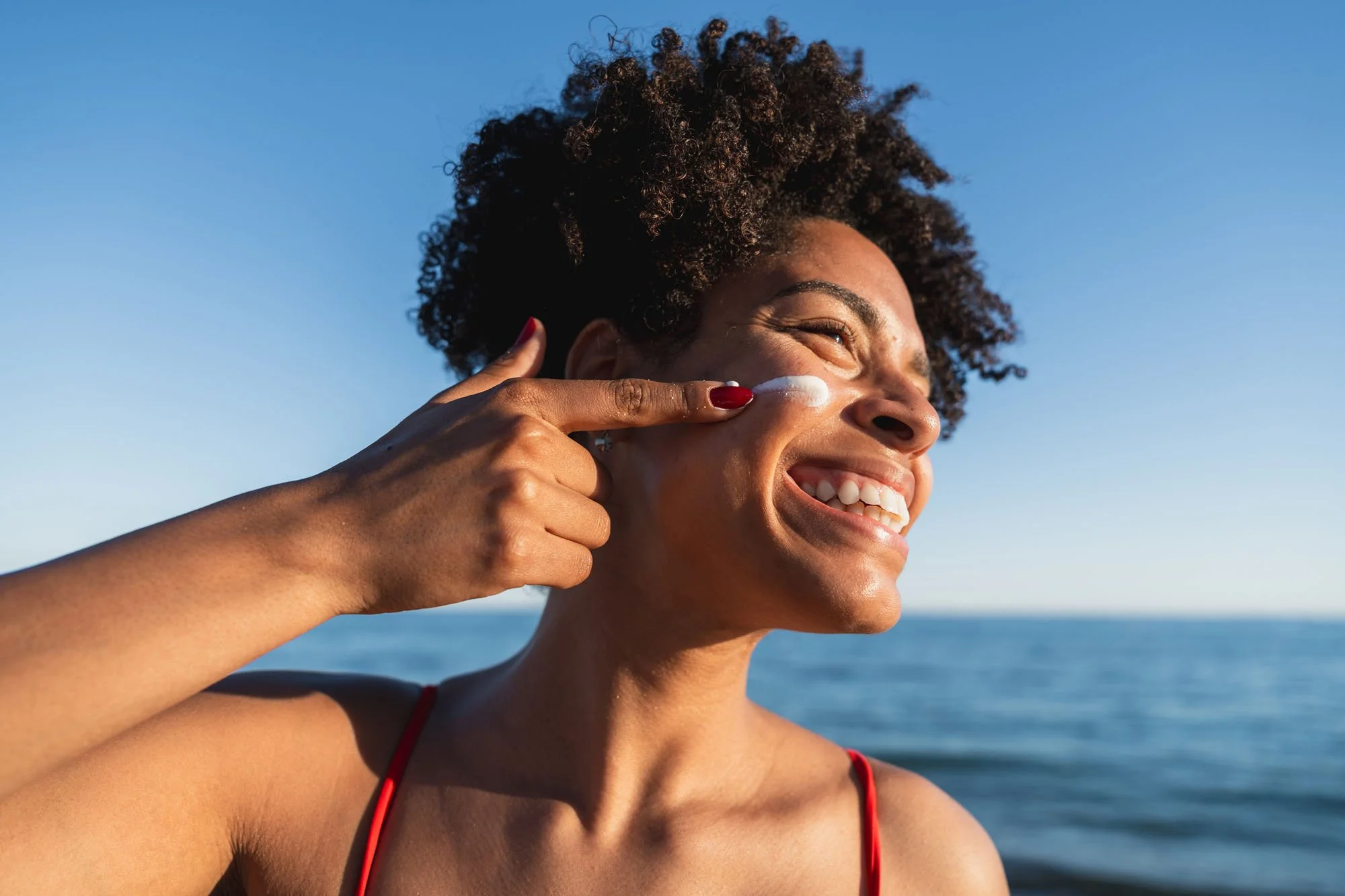 Mujer sonriendo en la playa, tocándose la mejilla con crema solar en el rostro, cabello rizado y oscuro, vestido de tirantes rojos, cielo despejado al fondo.