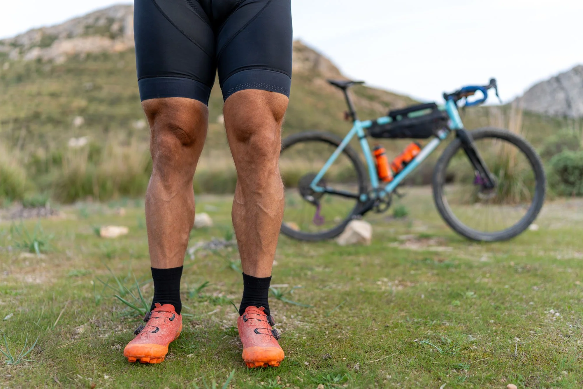Persona de espaldas con pantalones cortos de ciclismo, camiseta sin mangas y zapatillas deportivas, con una bicicleta de montaña en un paisaje natural.