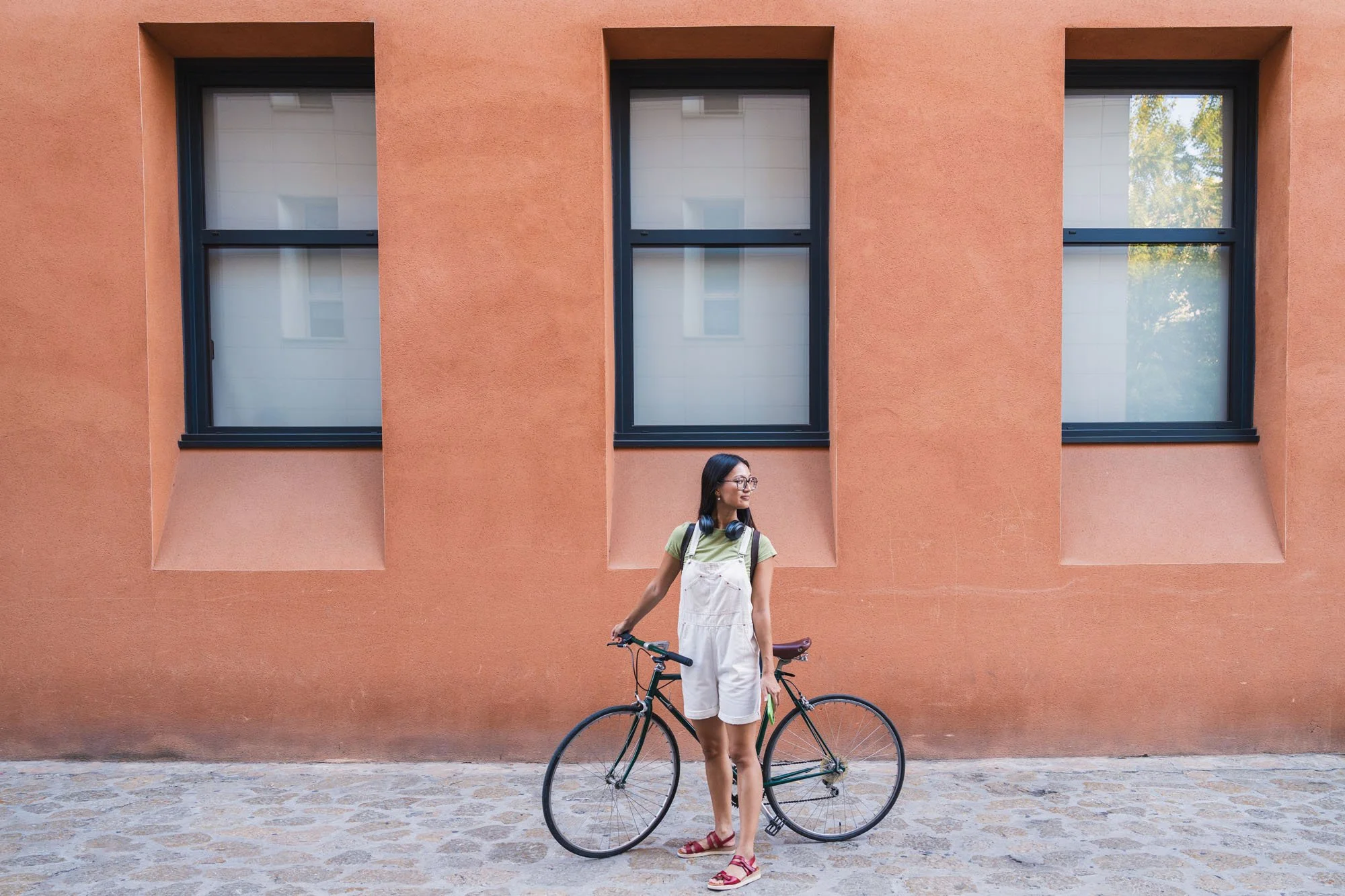 Joven mujer con gafas, camiseta verde, overol blanco y sandalias rojas, de pie con bicicleta frente a una pared naranja con ventanas negras.