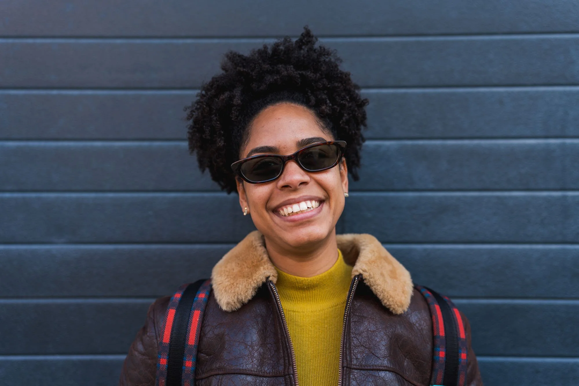 Mujer sonriente con gafas de sol oscuras, cabello rizado y oscuro, con chaqueta de cuero marrón con cuello de peluche, en un fondo de paredes grises horizontales.