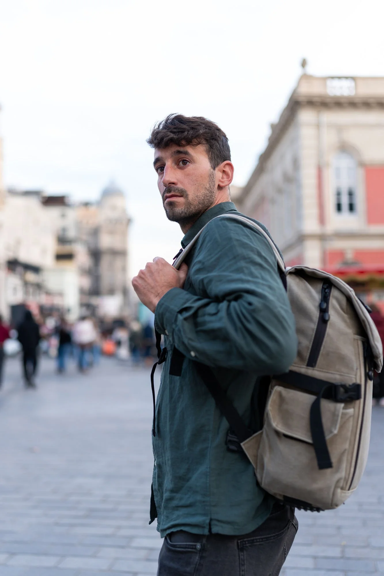 Joven con barba y cabello oscuro, con mochila beige, en una plaza urbana rodeado de edificios antiguos. Él mira hacia un lado, con expresión pensativa, mientras camina entre personas.