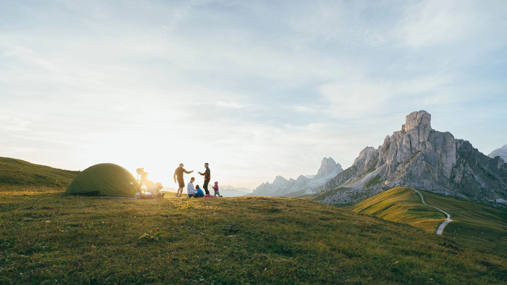 Un grupo de personas acampando en una pradera montañosa, con una tienda de campaña y montañas en el fondo, durante el atardecer.
