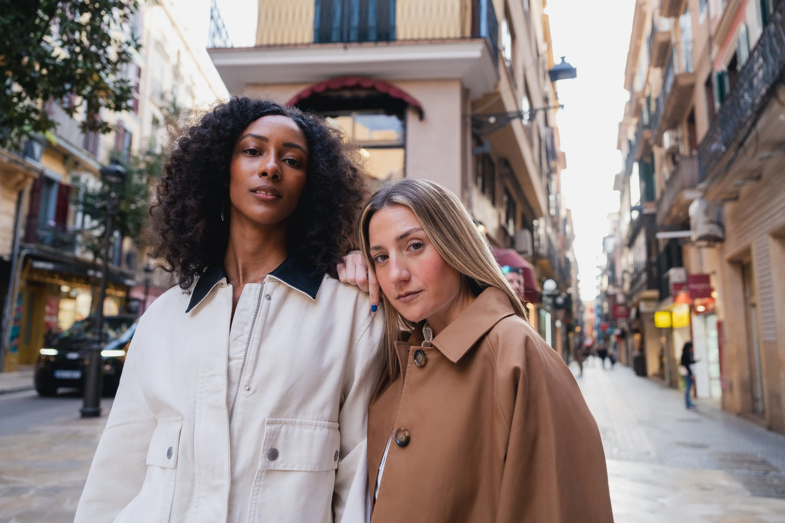 Dos mujeres jóvenes de pie juntas en una calle urbana, una con cabello rizado oscuro y piel oscura, la otra con cabello liso rubio y piel clara, ambas mirando hacia la cámara.