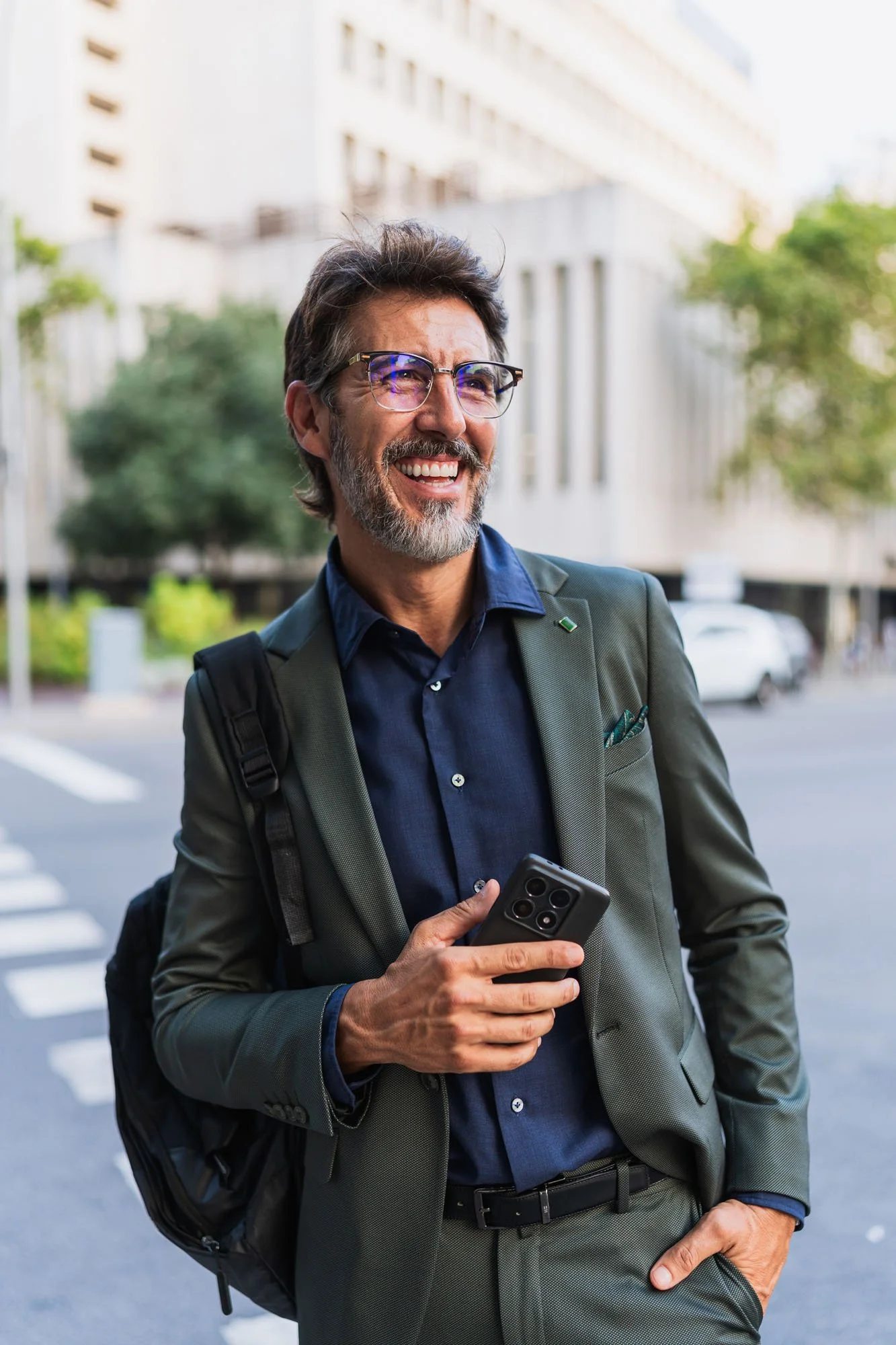 Hombre sonriente con barba y gafas, vestido con traje y camisa, sosteniendo un teléfono móvil, en una calle urbana con edificios y árboles.