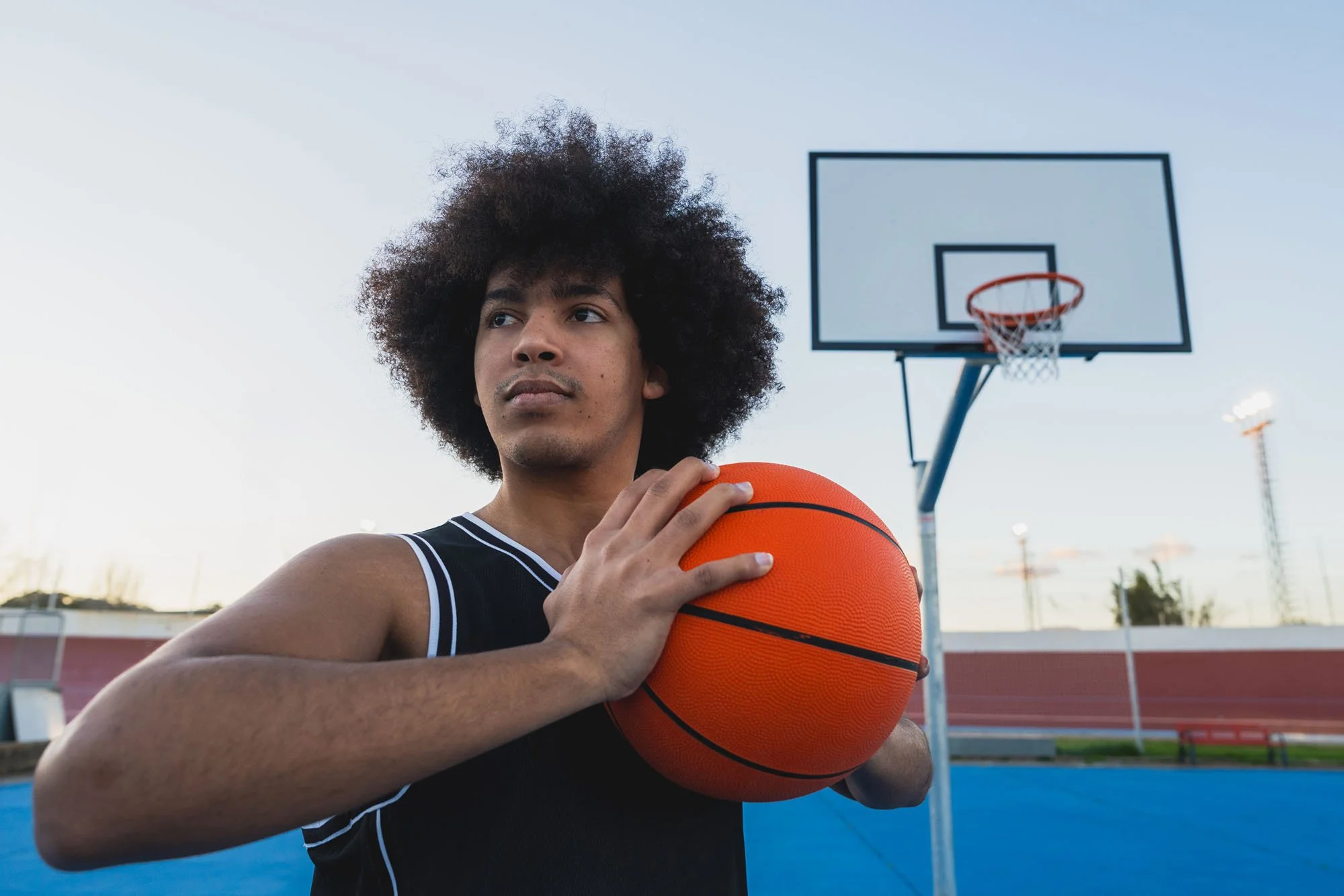 Joven jugador de baloncesto con uniforme negro, sosteniendo una pelota naranja en un parque de canchas de baloncesto al aire libre durante el atardecer.