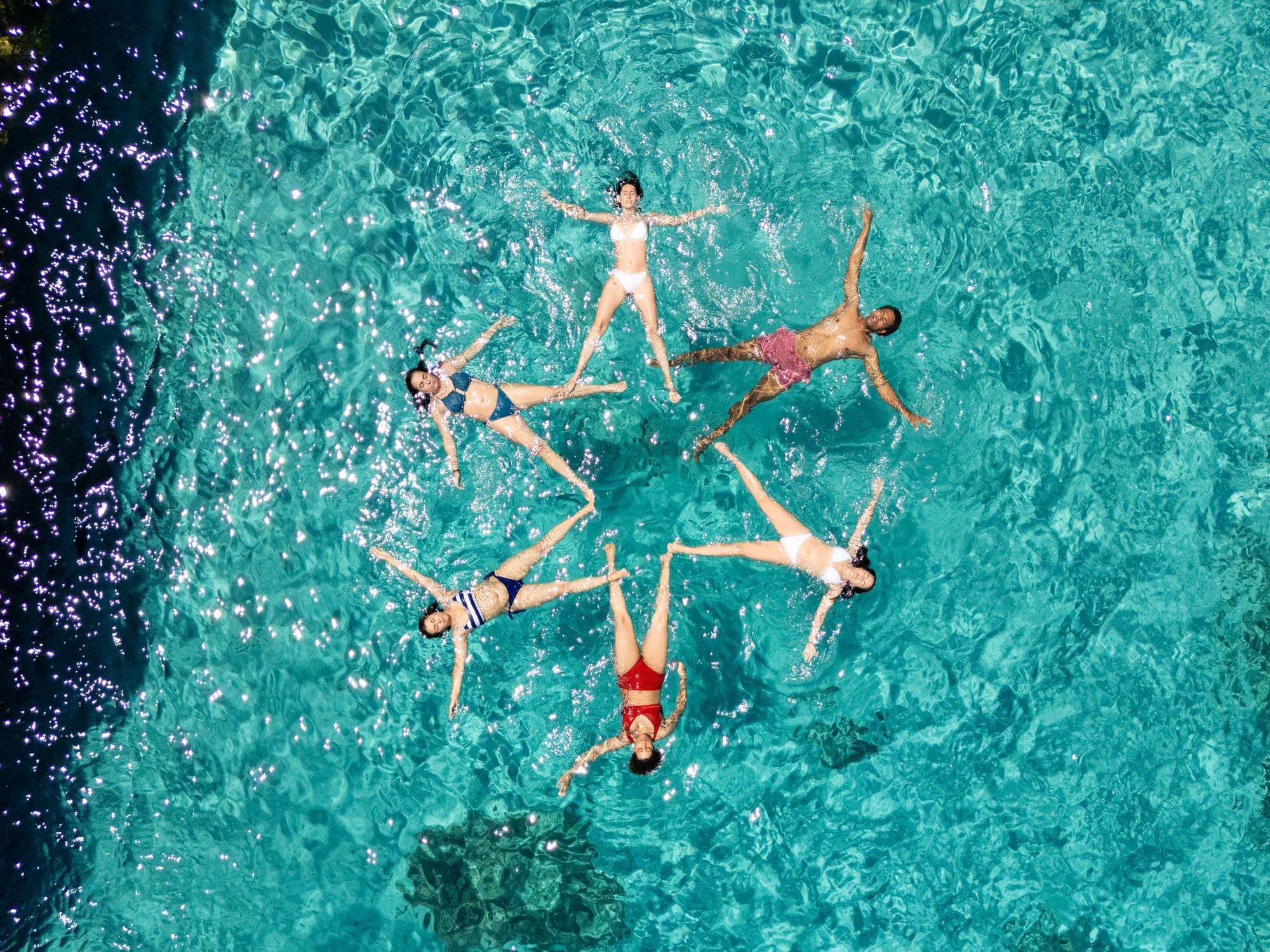 Diversas personas flotando en una piscina con agua azul clara, algunas con trajes de baño de diferentes colores.