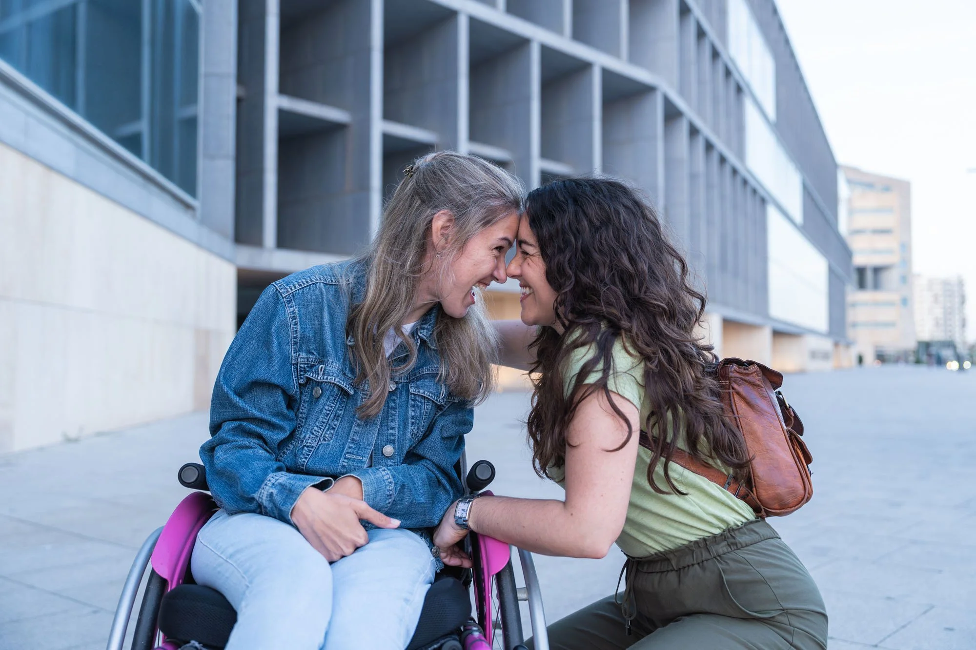 Dos mujeres jóvenes, una en silla de ruedas y otra apoyada en ella, comparten un momento feliz y cercano en un espacio exterior en una ciudad moderna, con edificios de fondo.