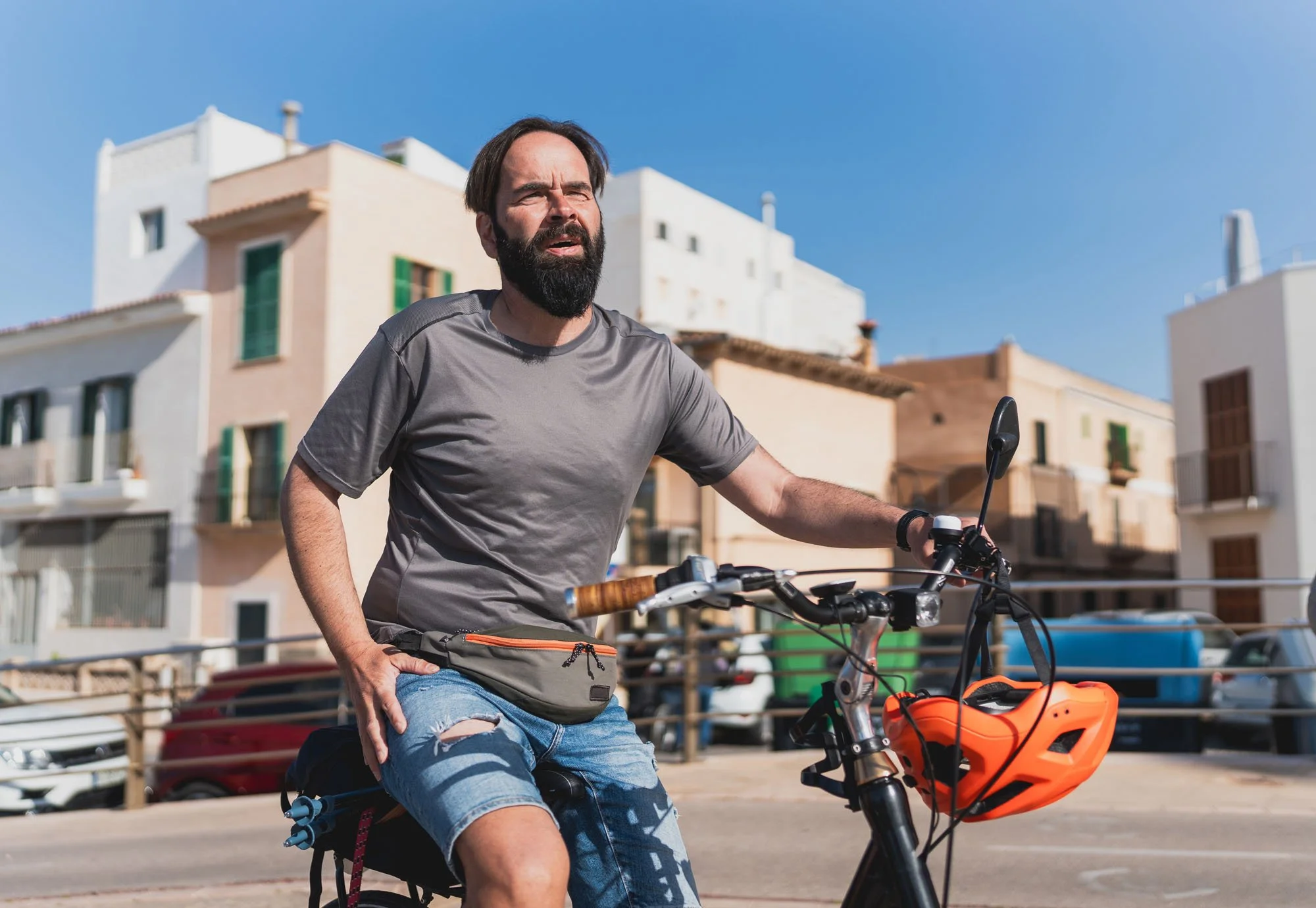Hombre con barba en bicicleta eléctrica en un callejón con edificios blancos al fondo bajo un cielo azul.