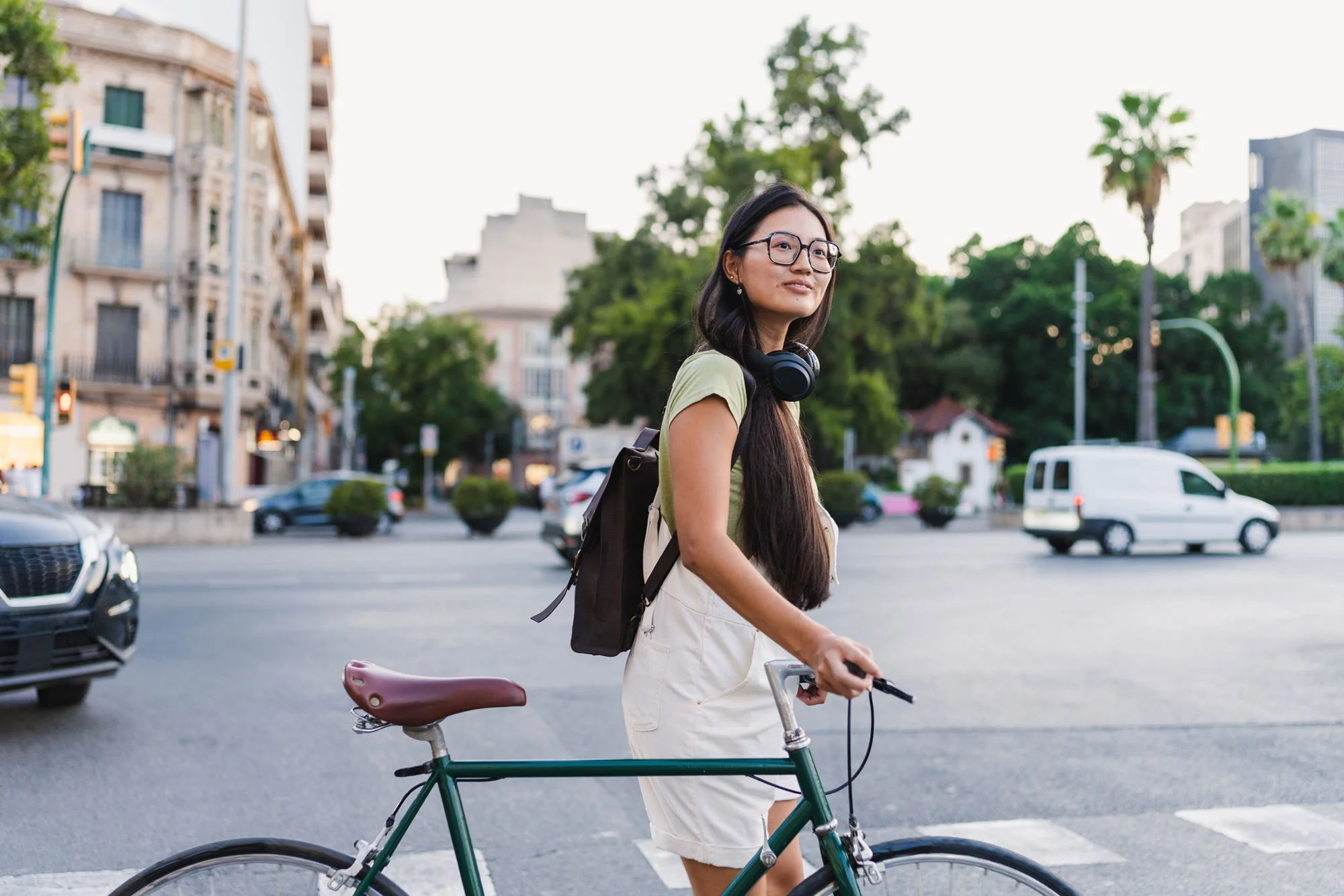 Mujer con gafas y auriculares caminando con bicicleta en la ciudad