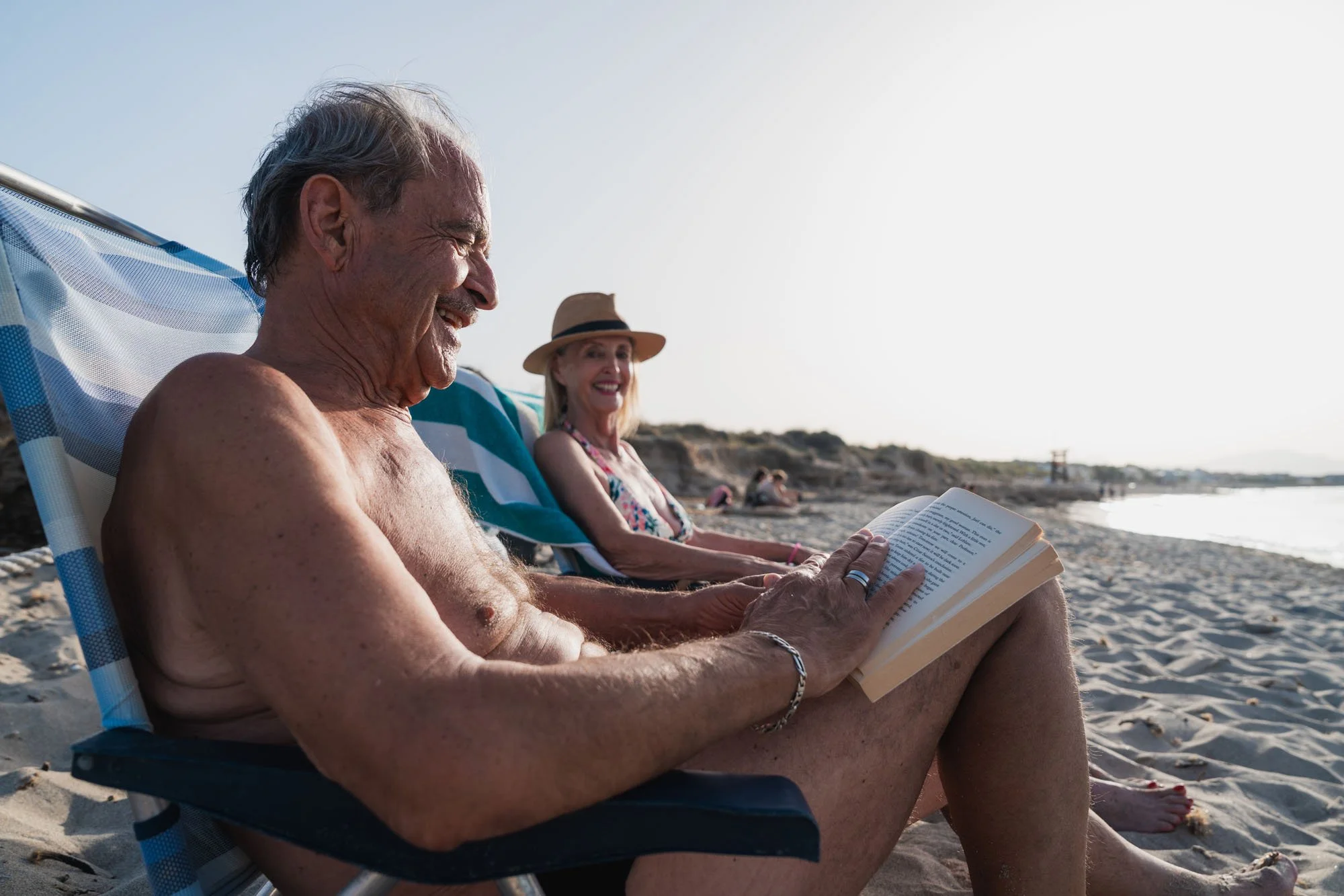 Un hombre mayor en una silla de playa leyendo un libro en la playa, acompañado de una mujer mayor que también disfruta del día soleado junto a la orilla del mar.