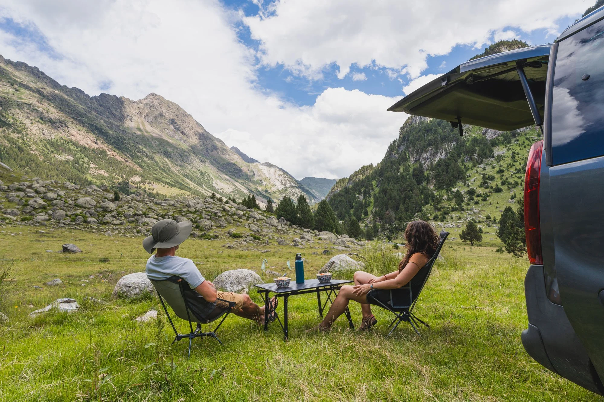 Dos personas en sillas de camping sentadas en un prado rodeado de montañas y árboles, con un techo de vehículo parcialmente visible a la derecha y un cielo con nubes.