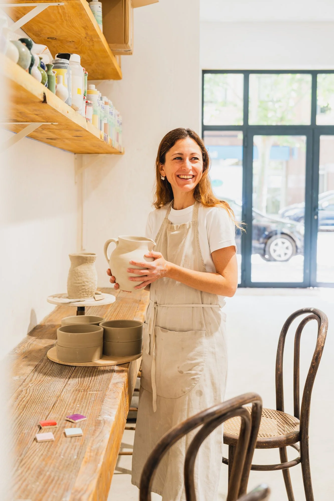 Mujer sonriendo en un taller de cerámica, sosteniendo una vasija. Hay estanterías con tazas y cerámicas a su lado, y varias verdaderas en proceso en la mesa de trabajo.
