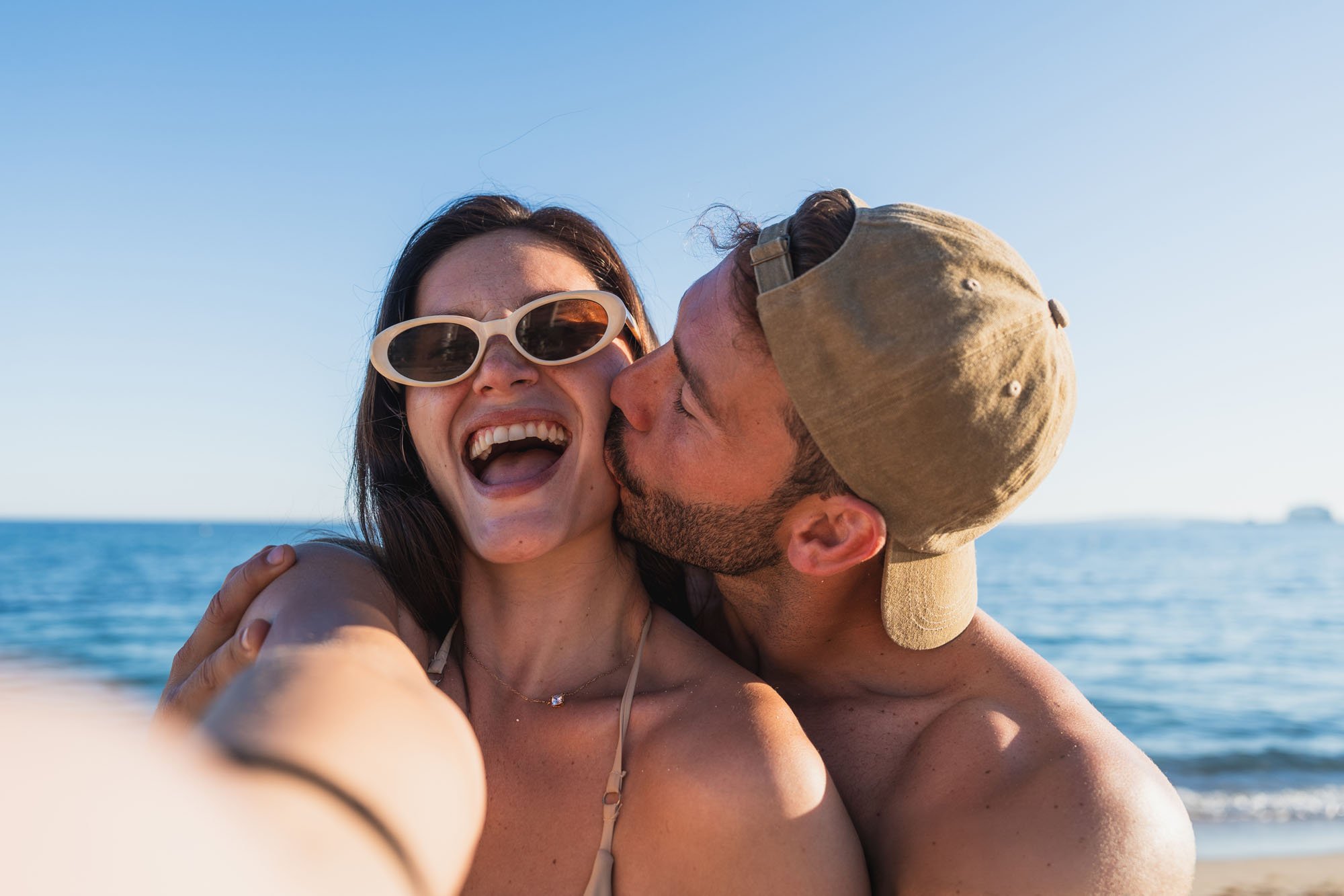 Pareja feliz en la playa tomándose una selfie, la mujer con gafas grandes y el hombre con gorra.