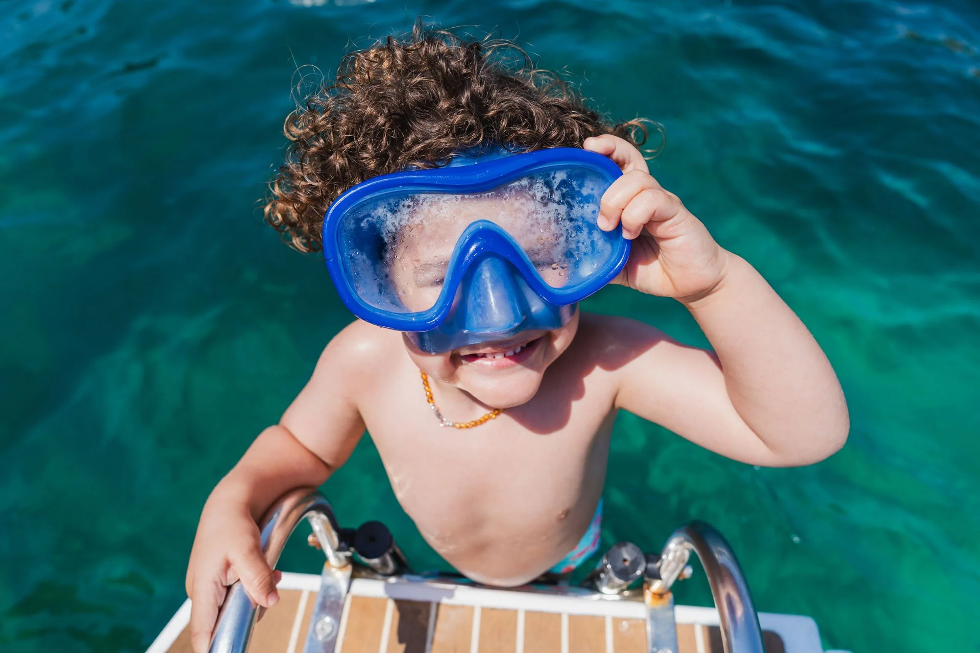 Niño con gafas de natación, sonriendo y disfrutando en el agua, mientras se sostiene de una estructura de metal en un entorno acuático.