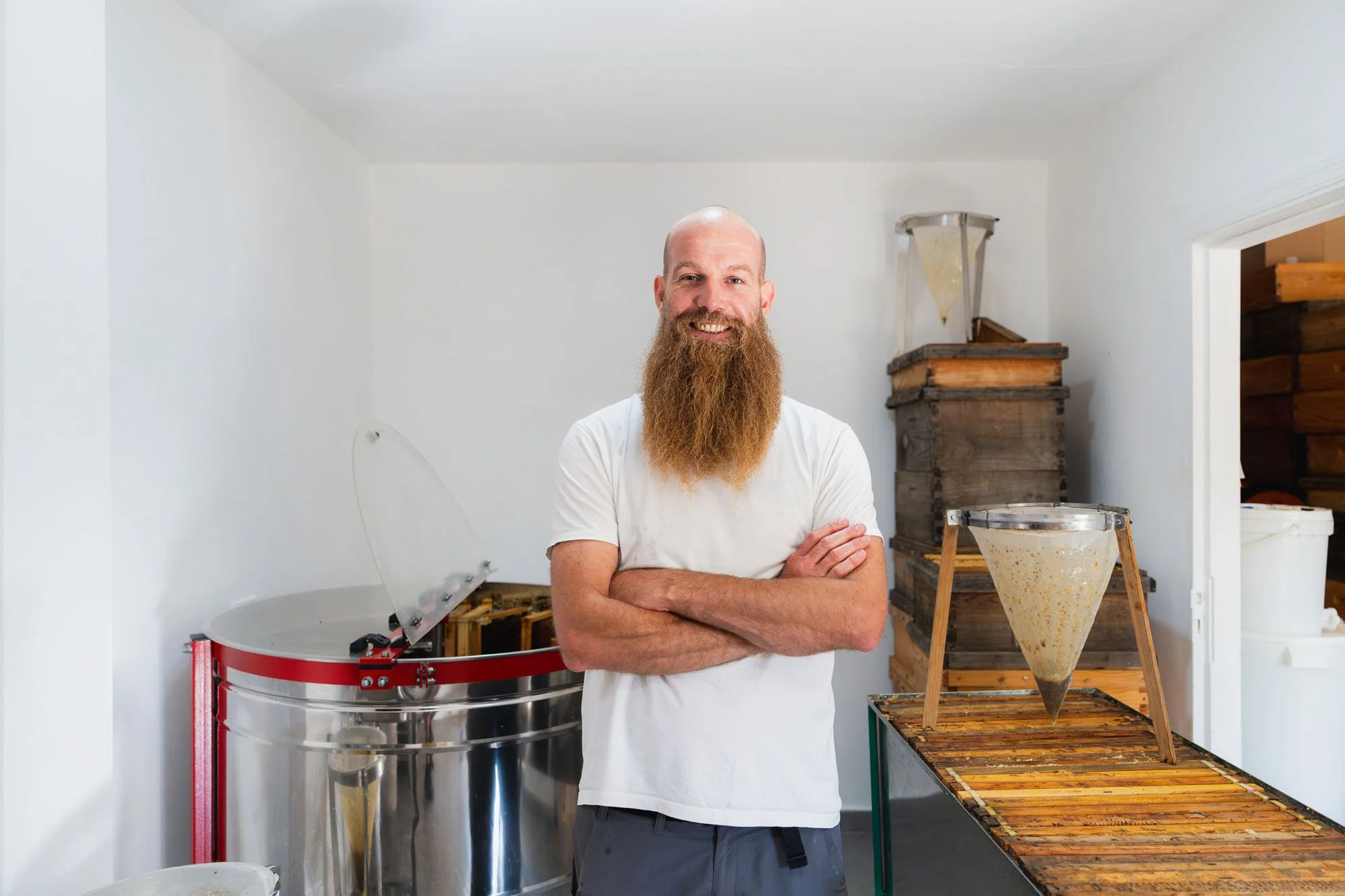 Hombre con barba rubia y camiseta blanca, sonriendo con brazos cruzados, en un taller con maquinaria y madera, probablemente haciendo producción artesanal.