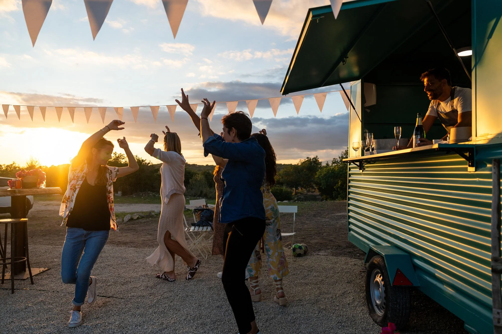 Varias personas bailan y disfrutan en un ambiente al aire libre con decoraciones de banderines, mientras un hombre en un puesto móvil de comida o bebidas sonríe y observa. El sol se está poniendo y el cielo está parcialmente nublado.