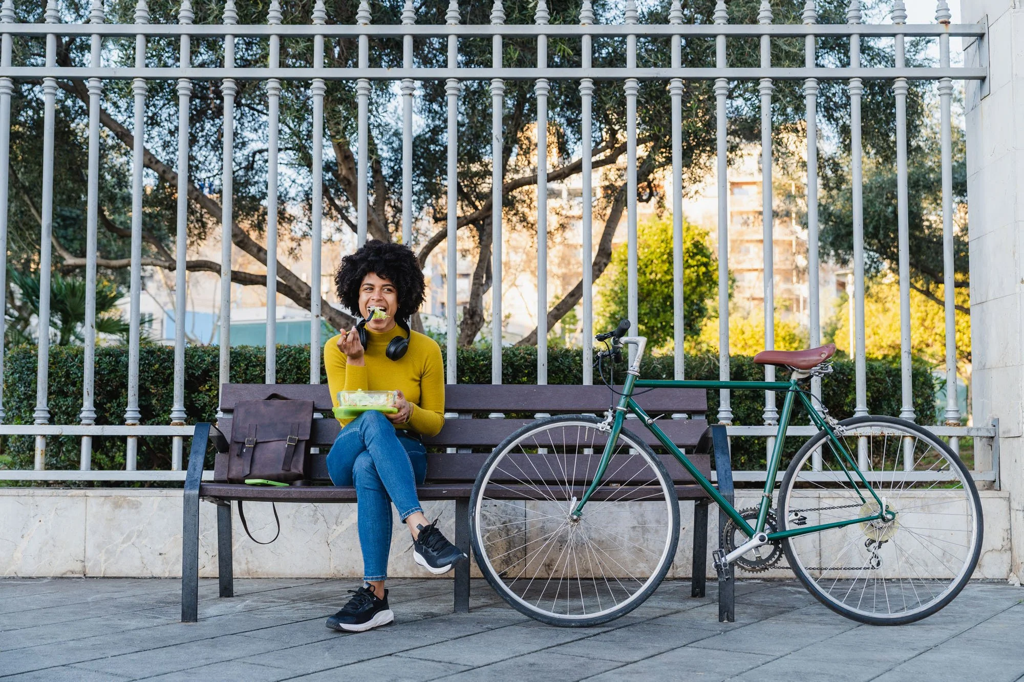 Mujer sentada en un banco comiendo ensalada, con bicicleta apoyada al banco, en un parque con árboles y una reja de metal al fondo.
