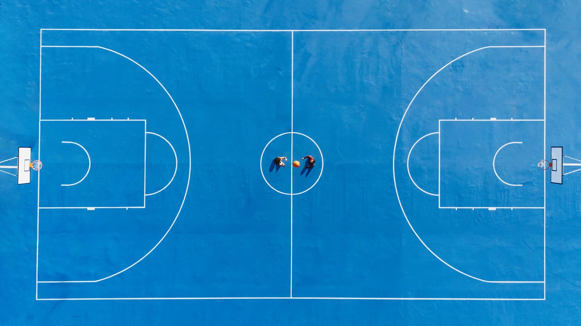 Vista aérea de una cancha de baloncesto azul con líneas blancas, dos personas en el centro con un balón de baloncesto