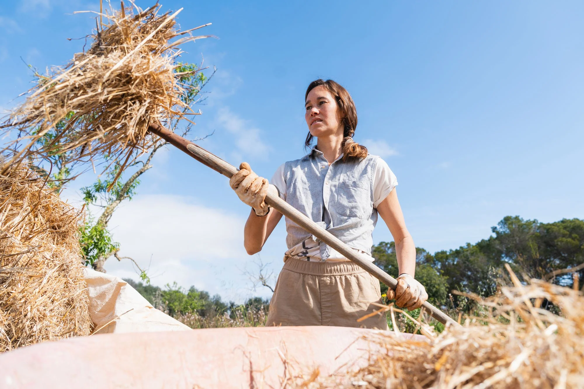 Mujer agricultora con guantes y uniforme trabaja con una herramienta en un campo con paja y vegetación en el fondo durante un día soleado.