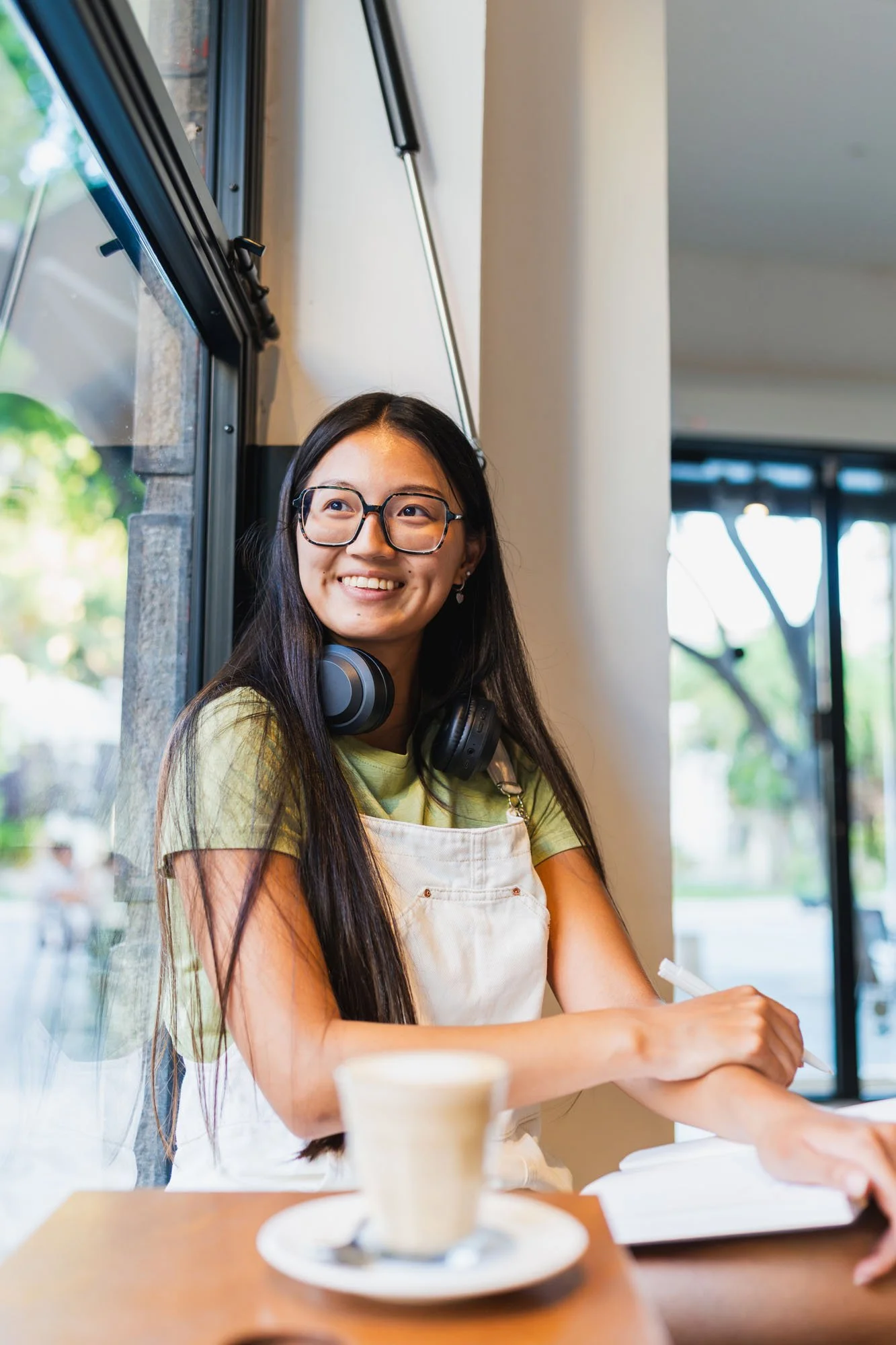 Joven mujer con gafas, auriculares y delantal, sonriendo junto a ventana, en ambiente interior, en una cafetería o panadería, mientras escribe en un cuaderno, con taza de café en primer plano.