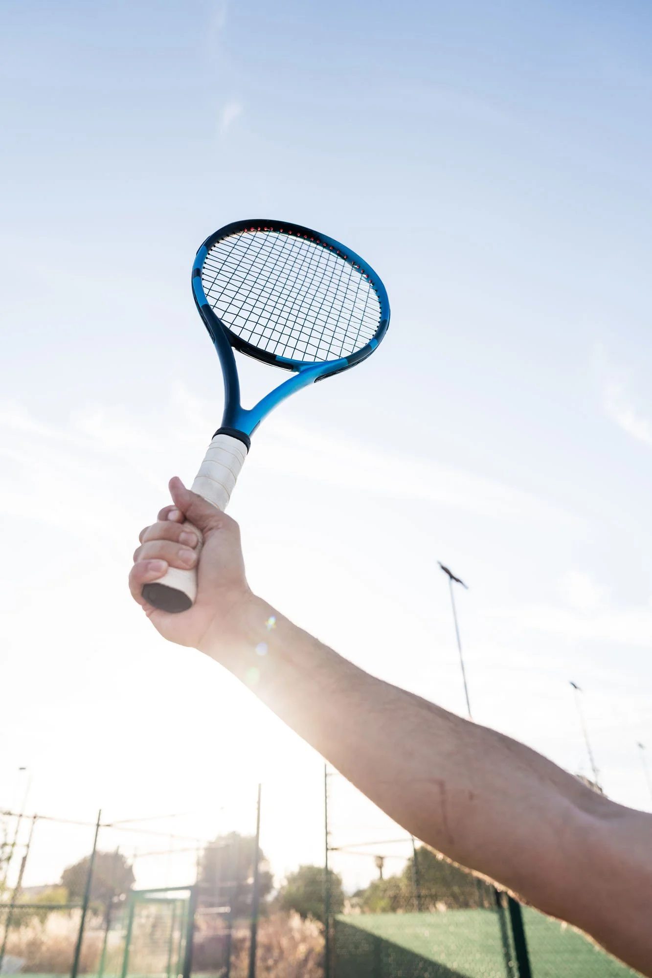 Una mano que sostiene una raqueta de tenis, apuntando hacia el cielo con un fondo de cielo claro y una cancha de tenis al fondo.