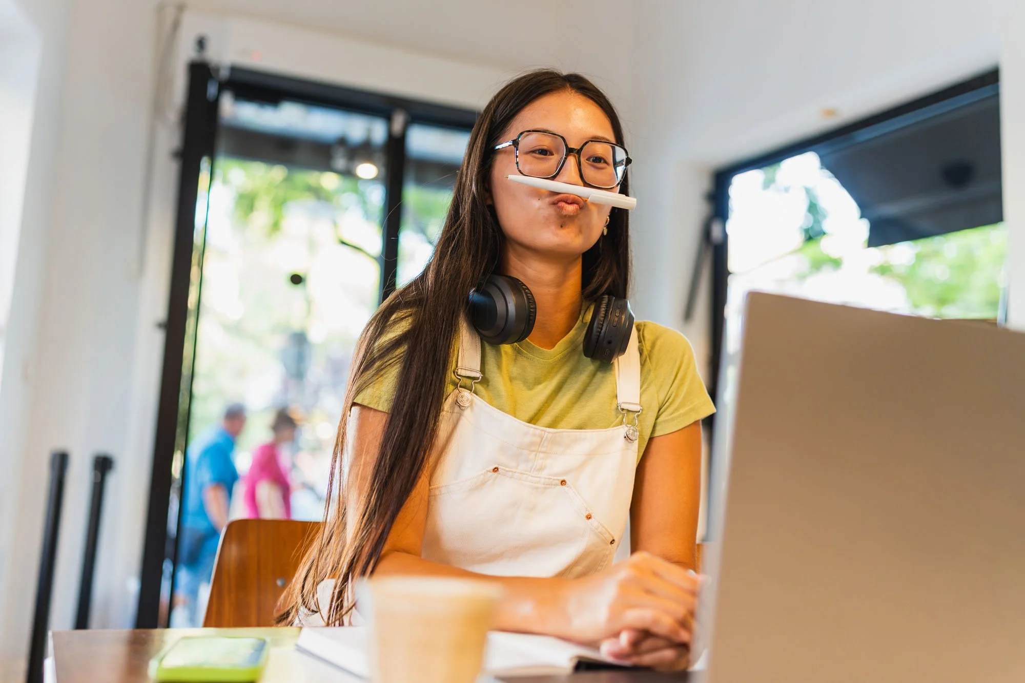 Joven mujer con gafas, con un bolígrafo en la boca, usando un delantal y camiseta verde, trabajando en una computadora en interiores bien iluminados con ventanas con vistas a árboles