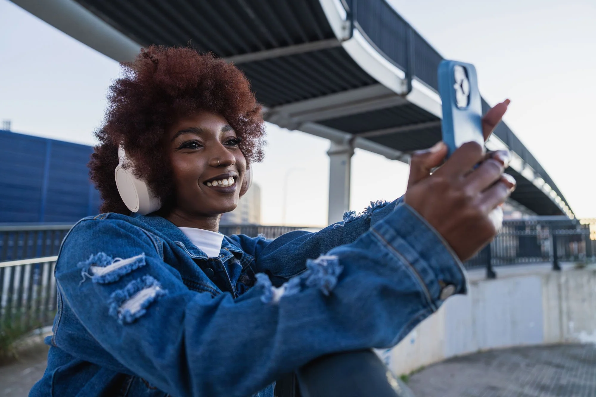 Juvenil sonriendo tomando una selfie con teléfono en una estación de tren o autobús con cielo despejado.