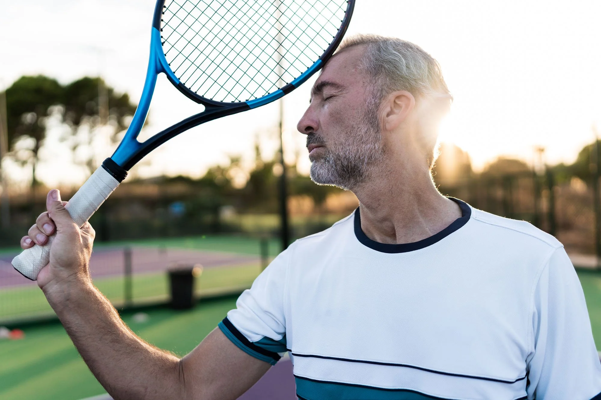 Hombre mayor con barba y cabello gris, con los ojos cerrados, sosteniendo una raqueta de tenis cerca de su cabeza en una cancha de tenis al aire libre mientras el sol se pone.