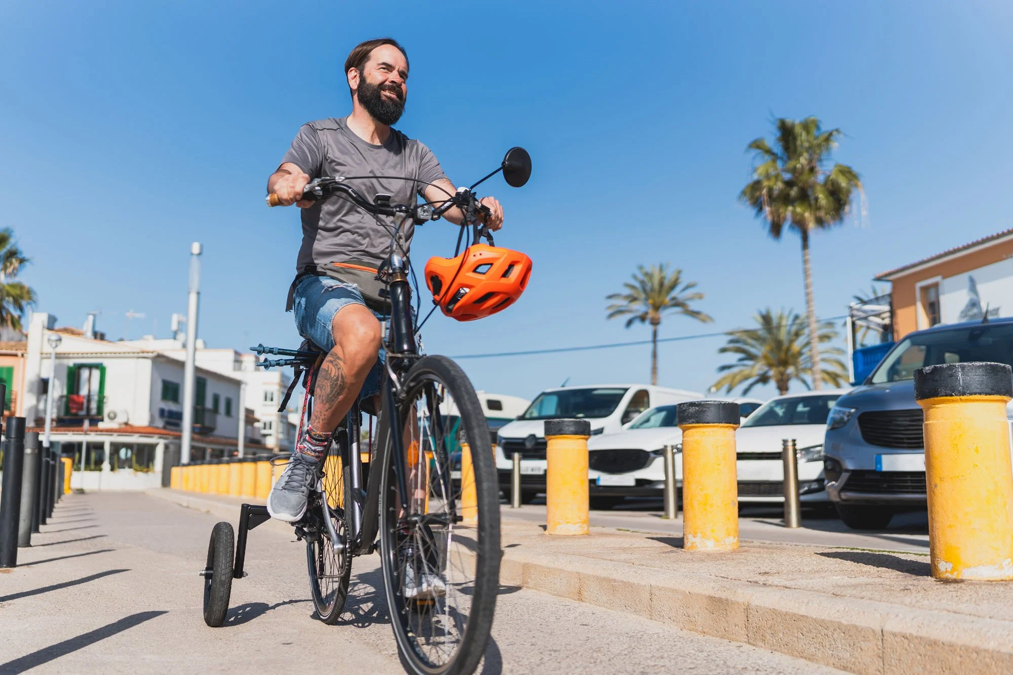 Un hombre con barba cabalga una bicicleta en un área urbana bajo un cielo despejado, con palmeras y edificios en el fondo, usa camiseta gris y pantalones cortos de mezclilla, lleva un casco naranja colgado en la bicicleta.