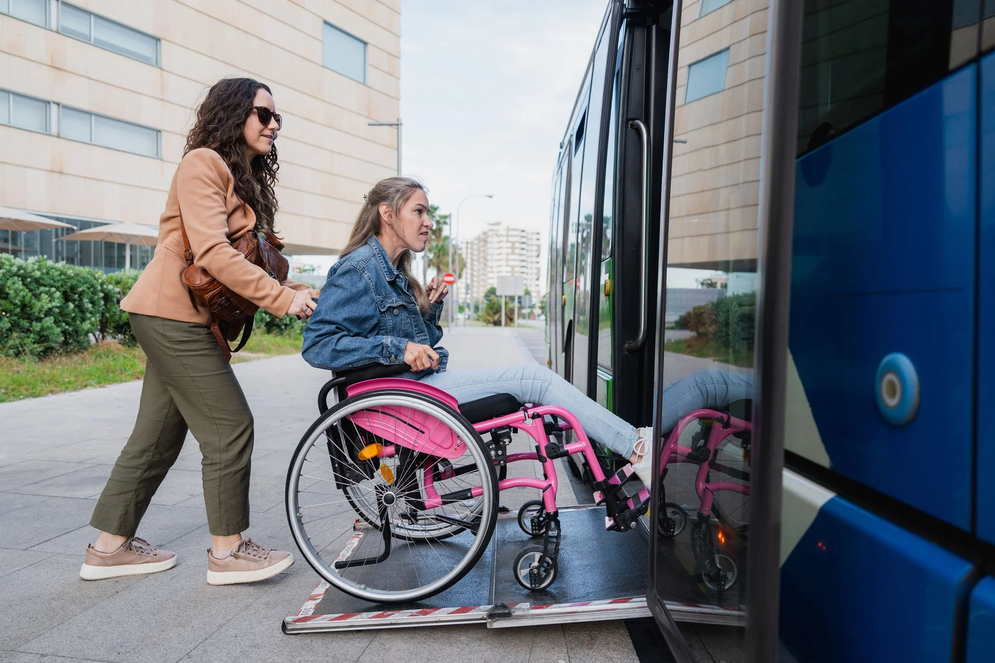 Una mujer en silla de ruedas y otra mujer están cargando alguien en una silla de ruedas a un autobús en un entorno urbano, con edificios modernos y árboles en el fondo.