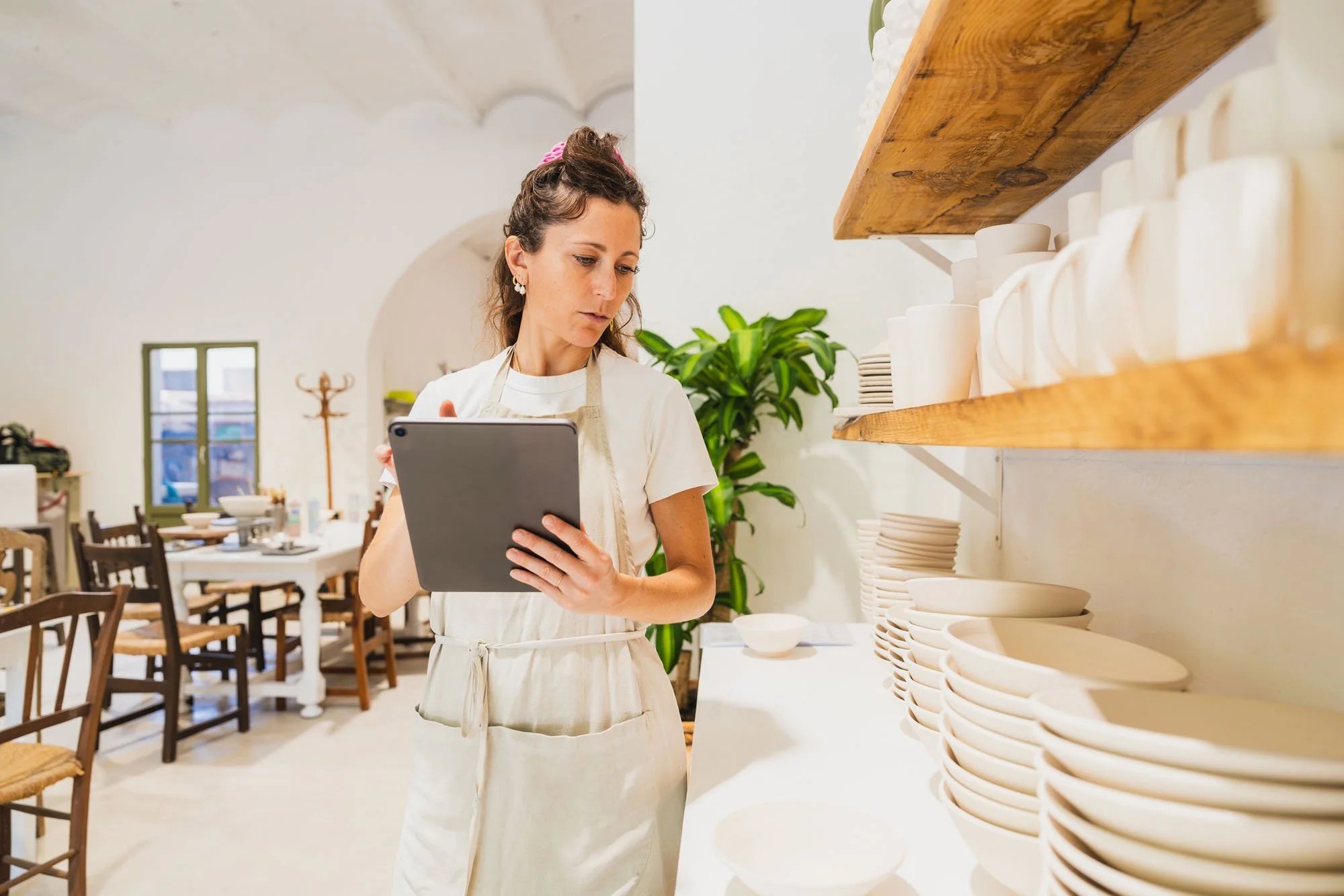 Mujer en del restaurante revisando una tableta, rodeada de platos blancos y estanterías con platos en la pared, en un ambiente con decoraciones sencillas y mucha luz natural.