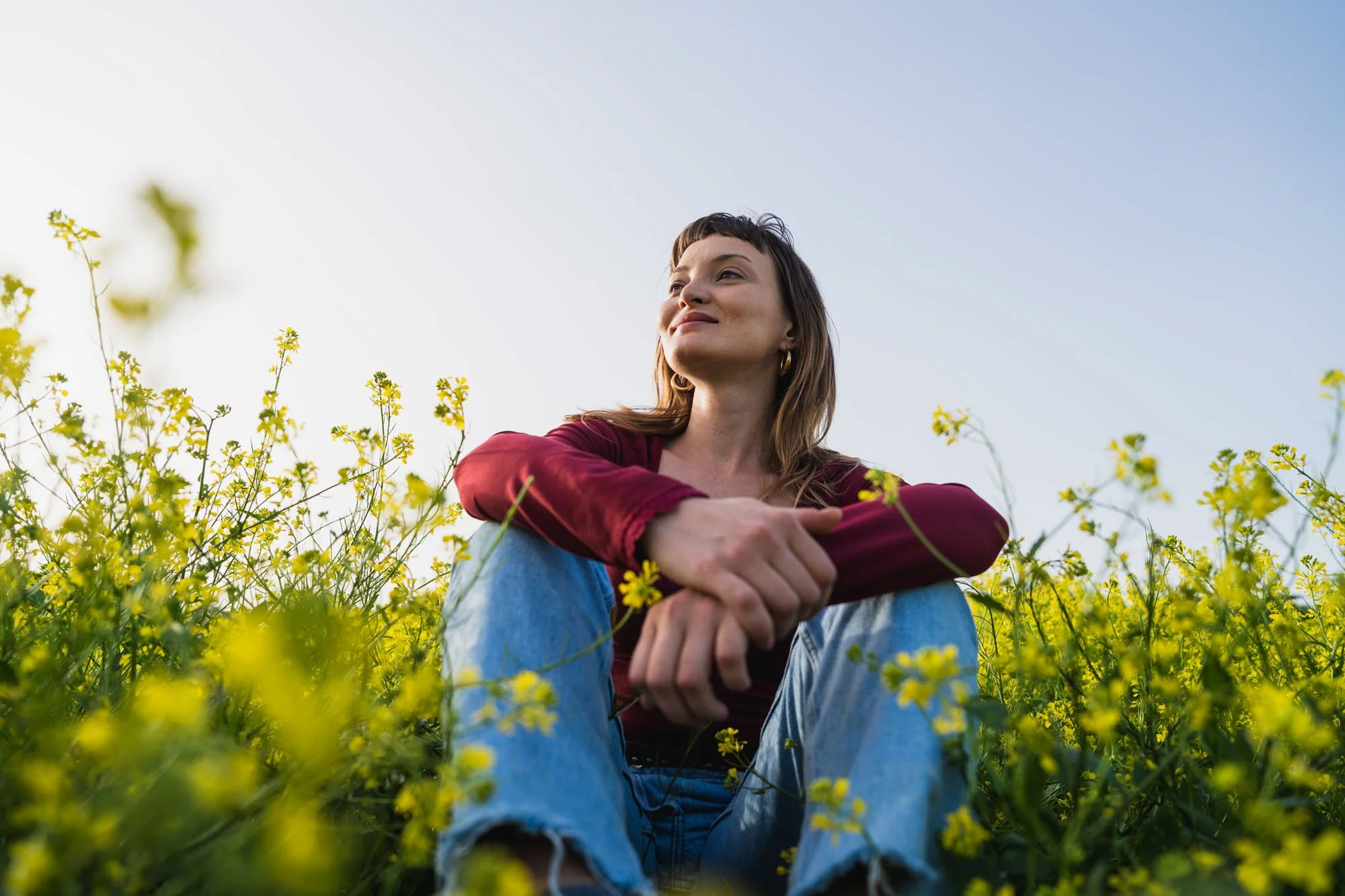 Mujer sentada entre flores amarillas con brazos cruzados, rostro tranquilo, mira hacia arriba, cielo despejado.